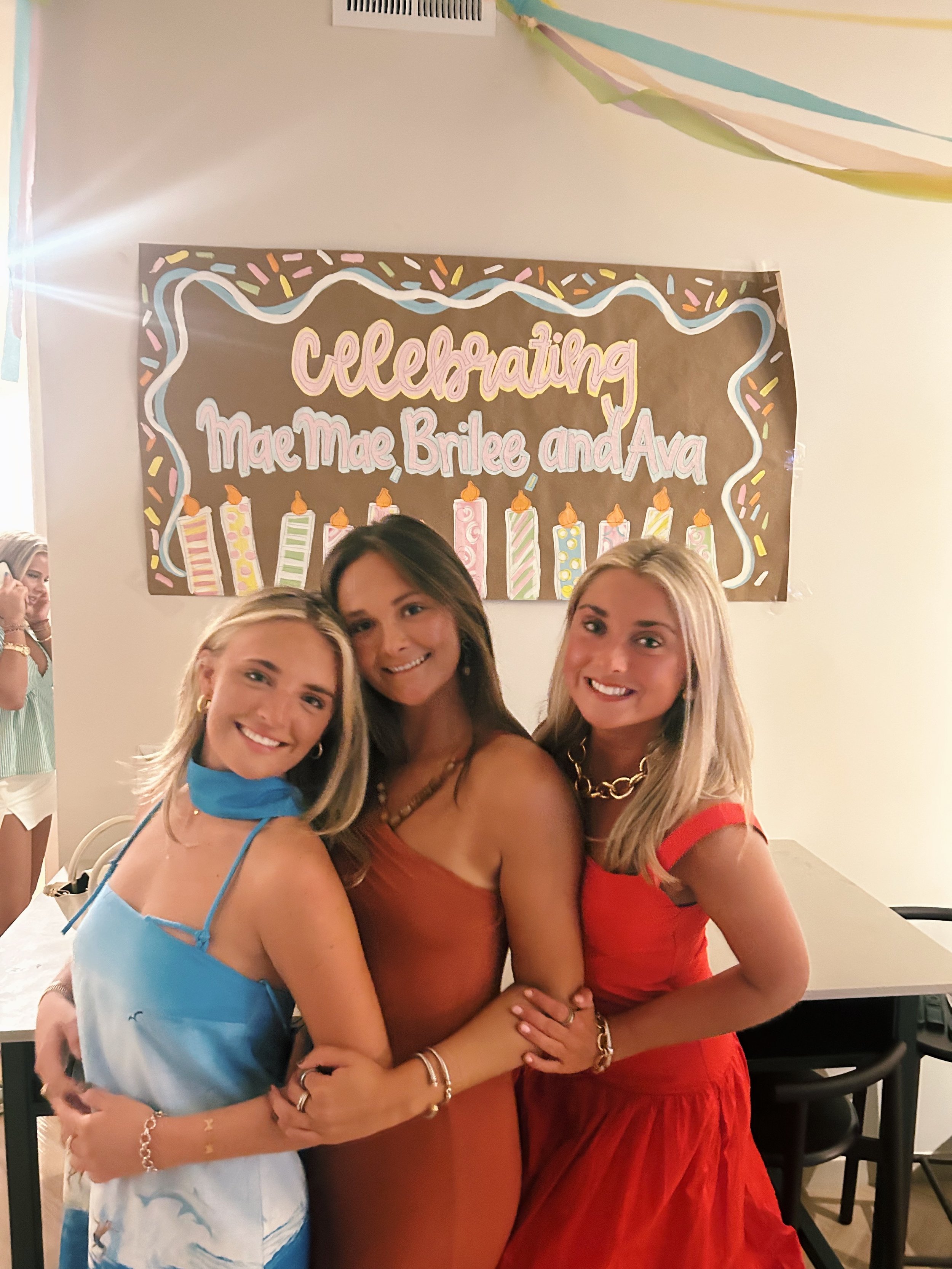 Three young women smiling and hugging in front of a birthday party banner that reads 'Celebrating Mae Mae, Brilee, and Ava' with colorful candles and sprinkles.