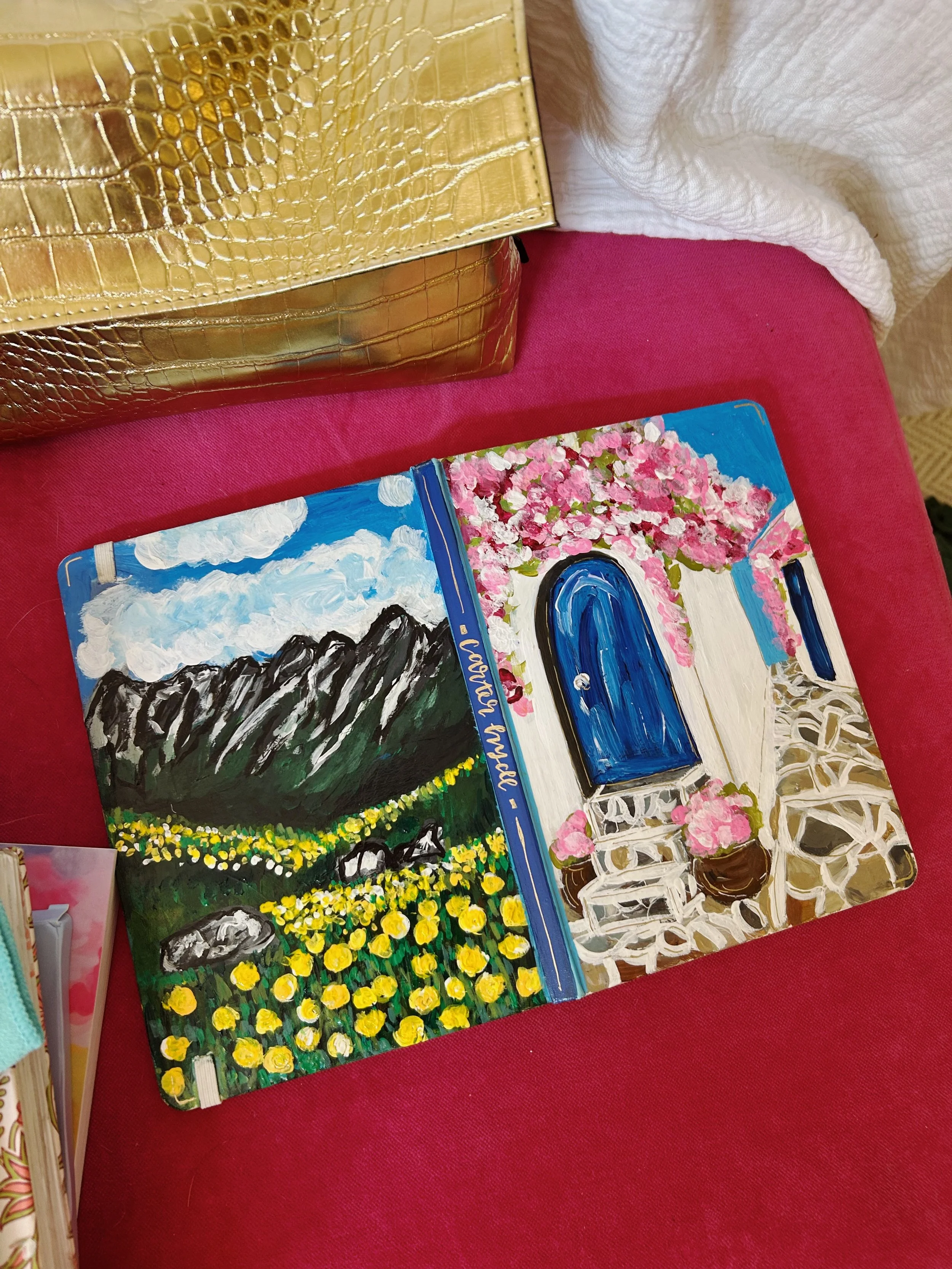 Painted journal with a landscape scene of mountains and yellow flowers on the left page, and a courtyard with a blue door and pink flowering trees on the right page, placed on a red surface next to a gold handbag.
