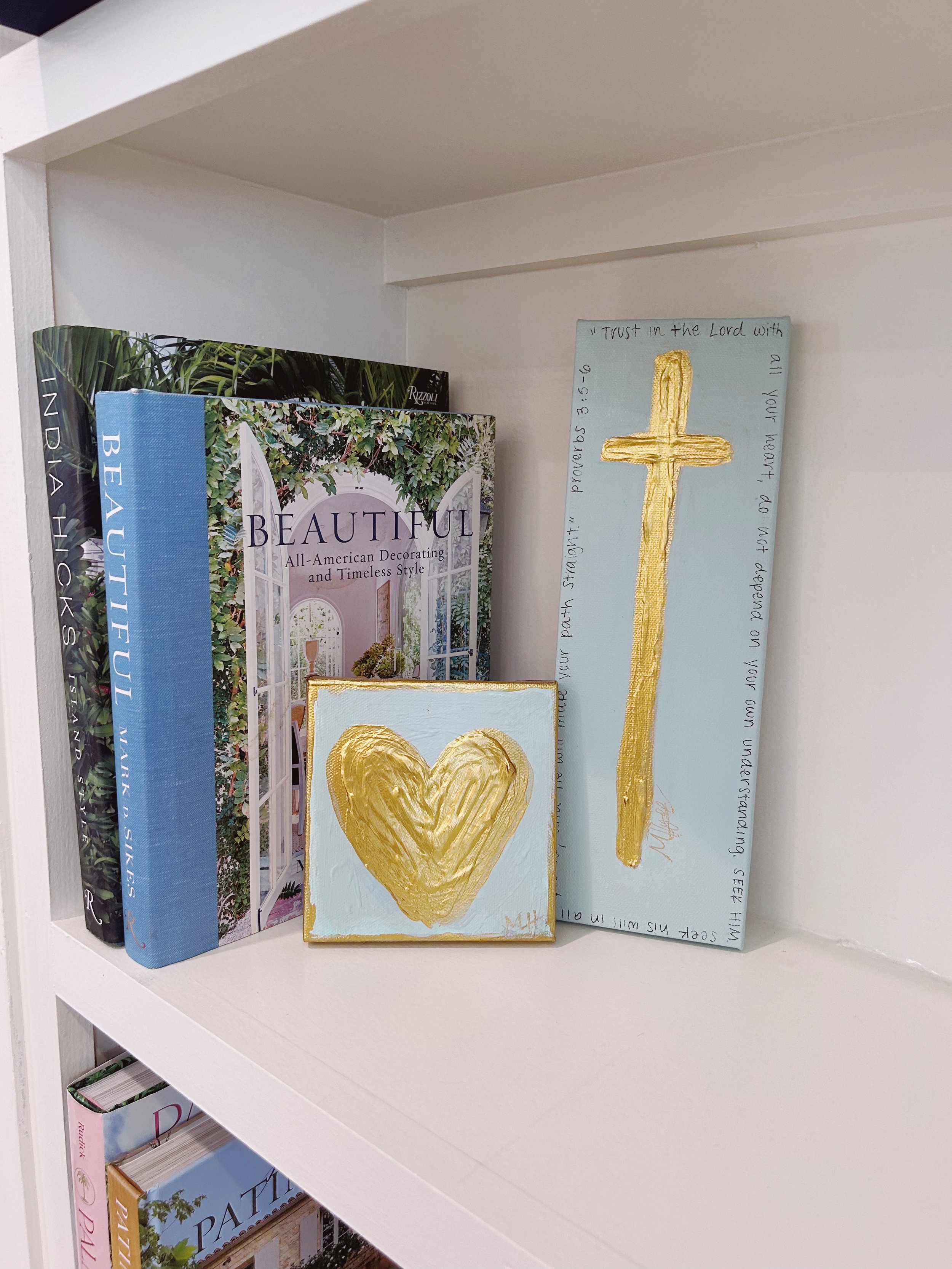 Decorative items on a white bookshelf, including a large book titled 'BEAUTIFUL', a smaller artwork with a gold heart, and a tall artwork with a gold cross.