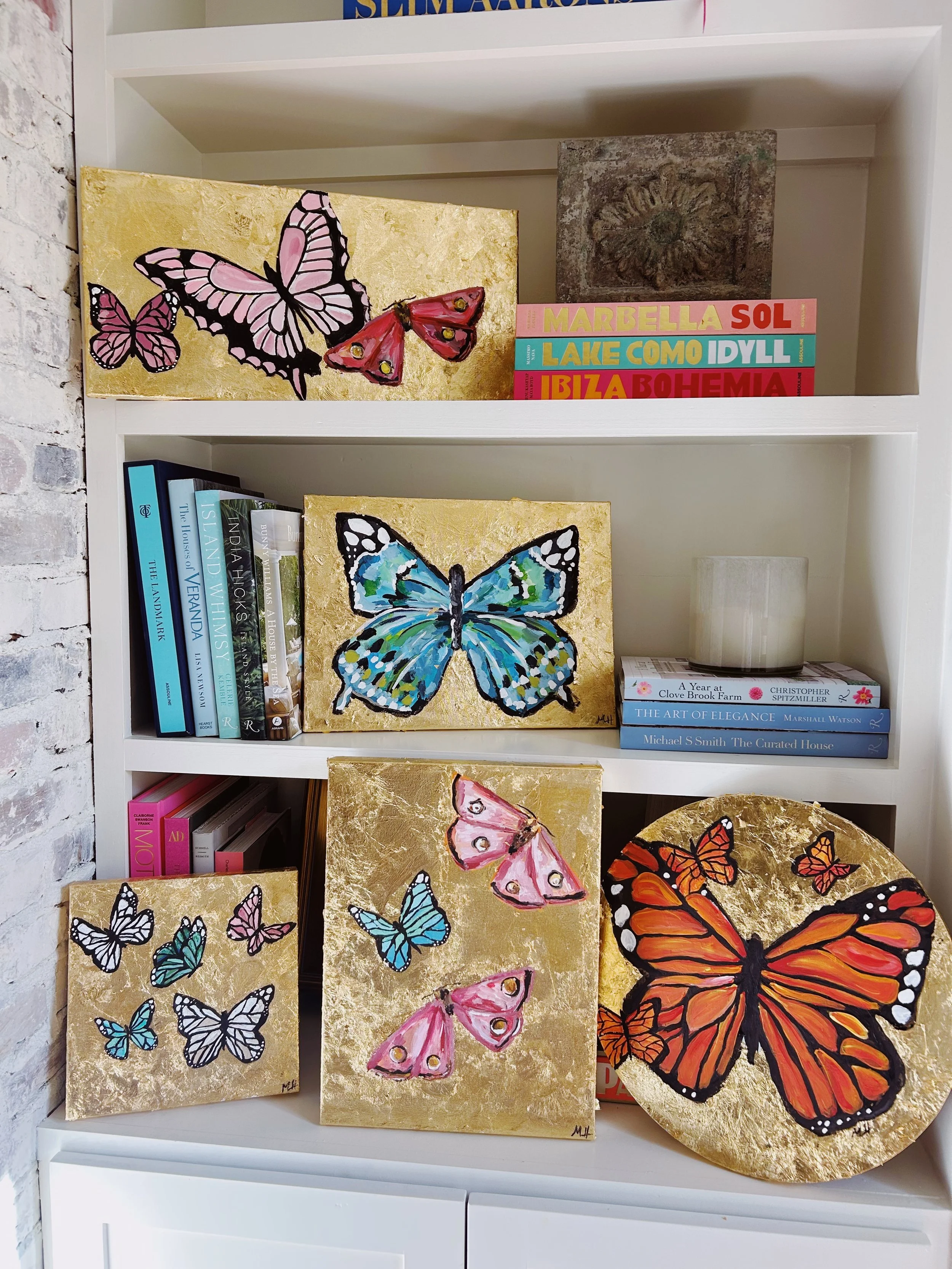 Decorative paintings of colorful butterflies arranged on a white bookshelf, with books and small decorative items around them.