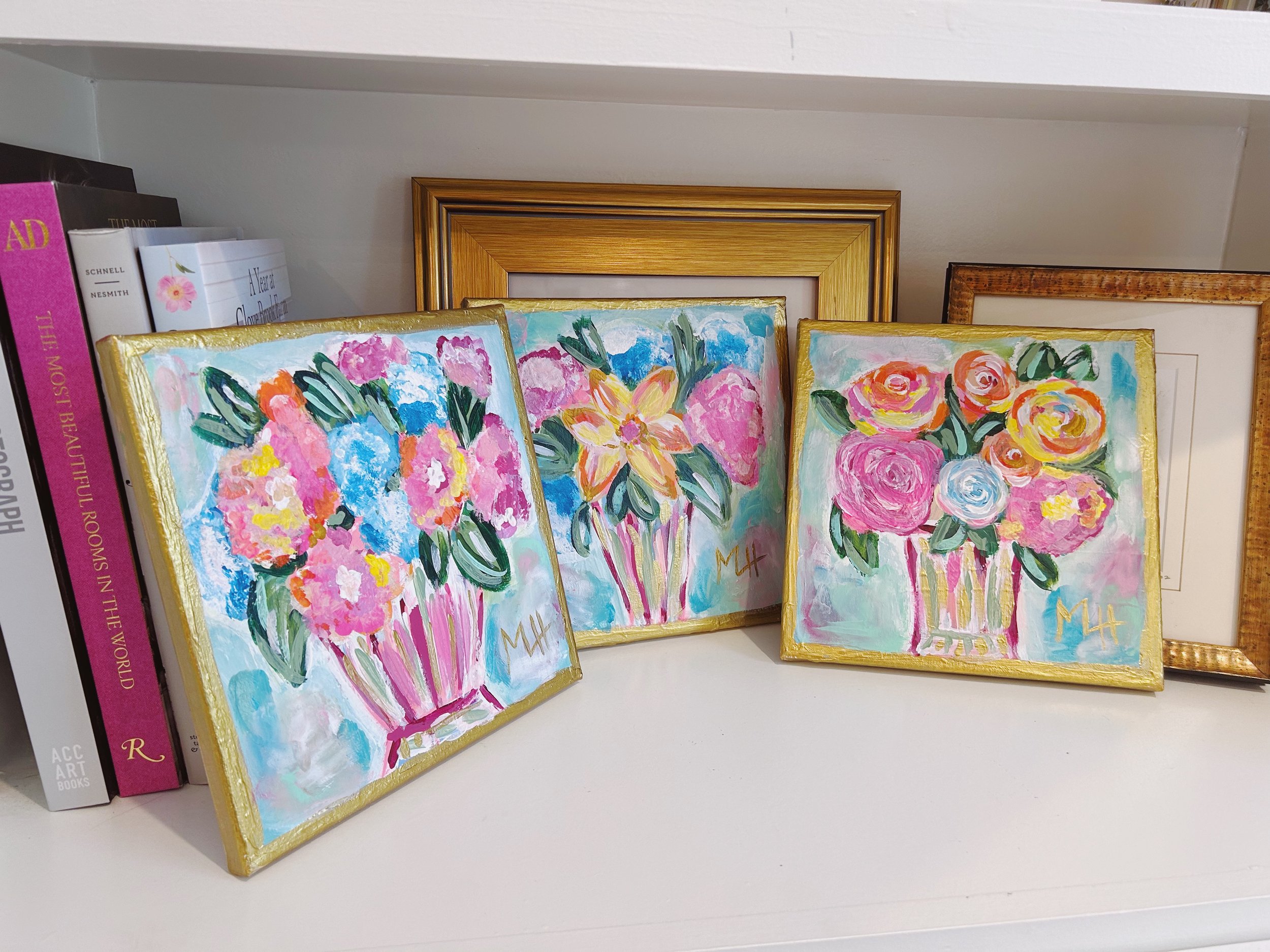 Three small colorful floral paintings in gold frames placed on a white shelf, next to books and larger framed pictures.