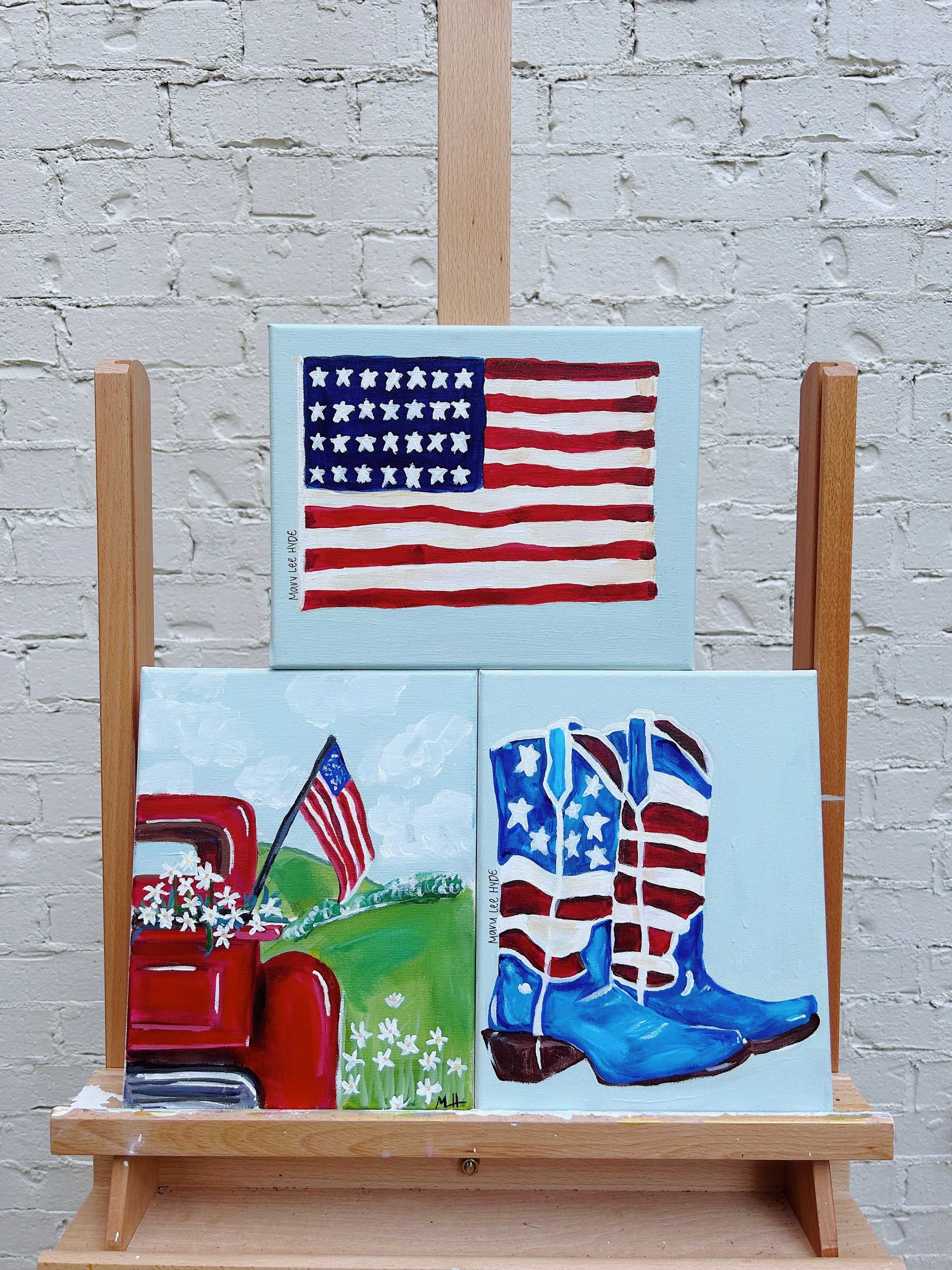 Three American-themed paintings on a wooden display stand against a white brick wall. The top painting features the American flag with blue field of stars and red and white stripes. The bottom left painting depicts a red truck with an American flag a