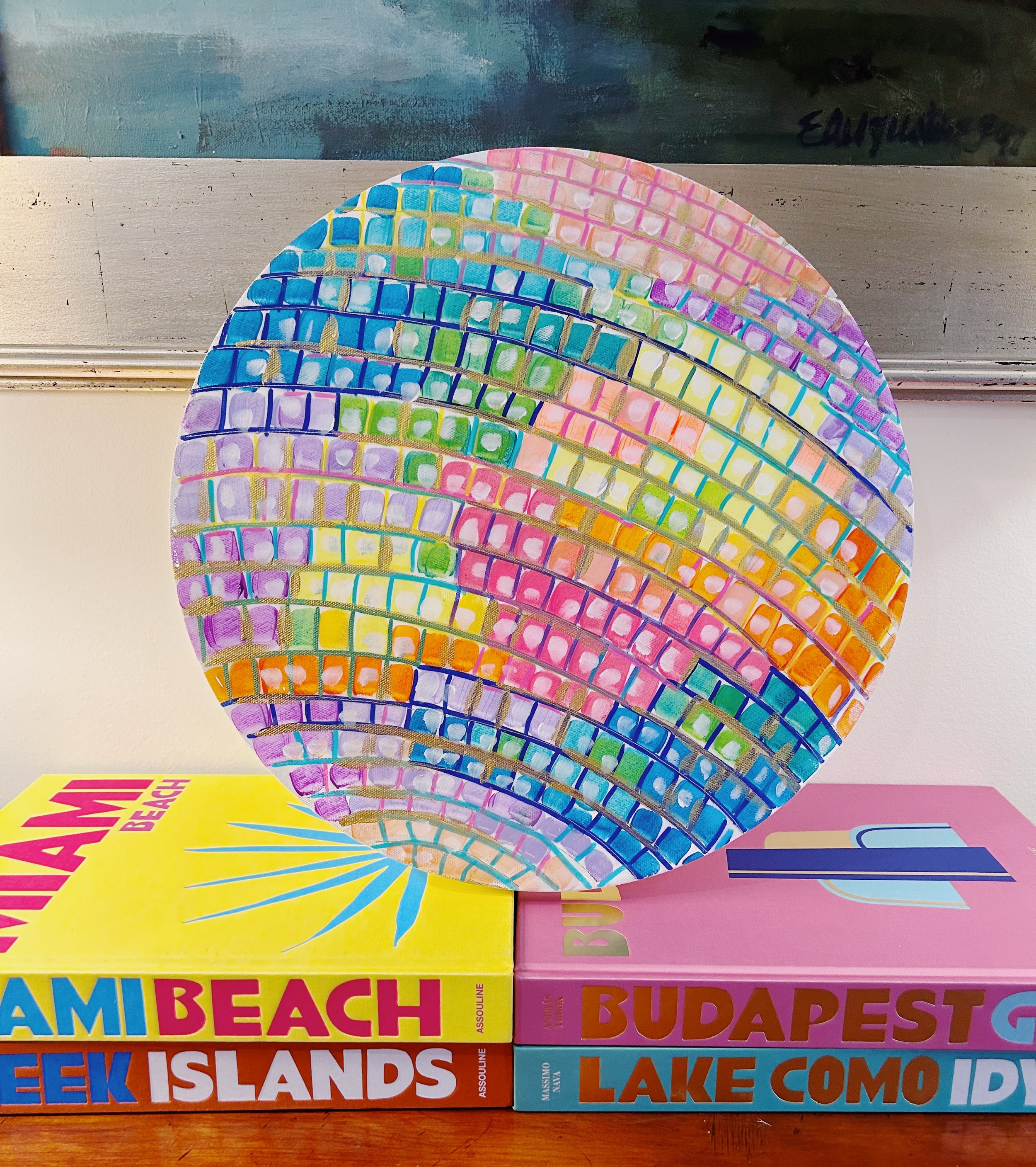 Colorful, abstract circular painting with a grid pattern of various pastel colors, placed on top of a stack of colorful travel books about Miami Beach, Budapest, and Lake Como.