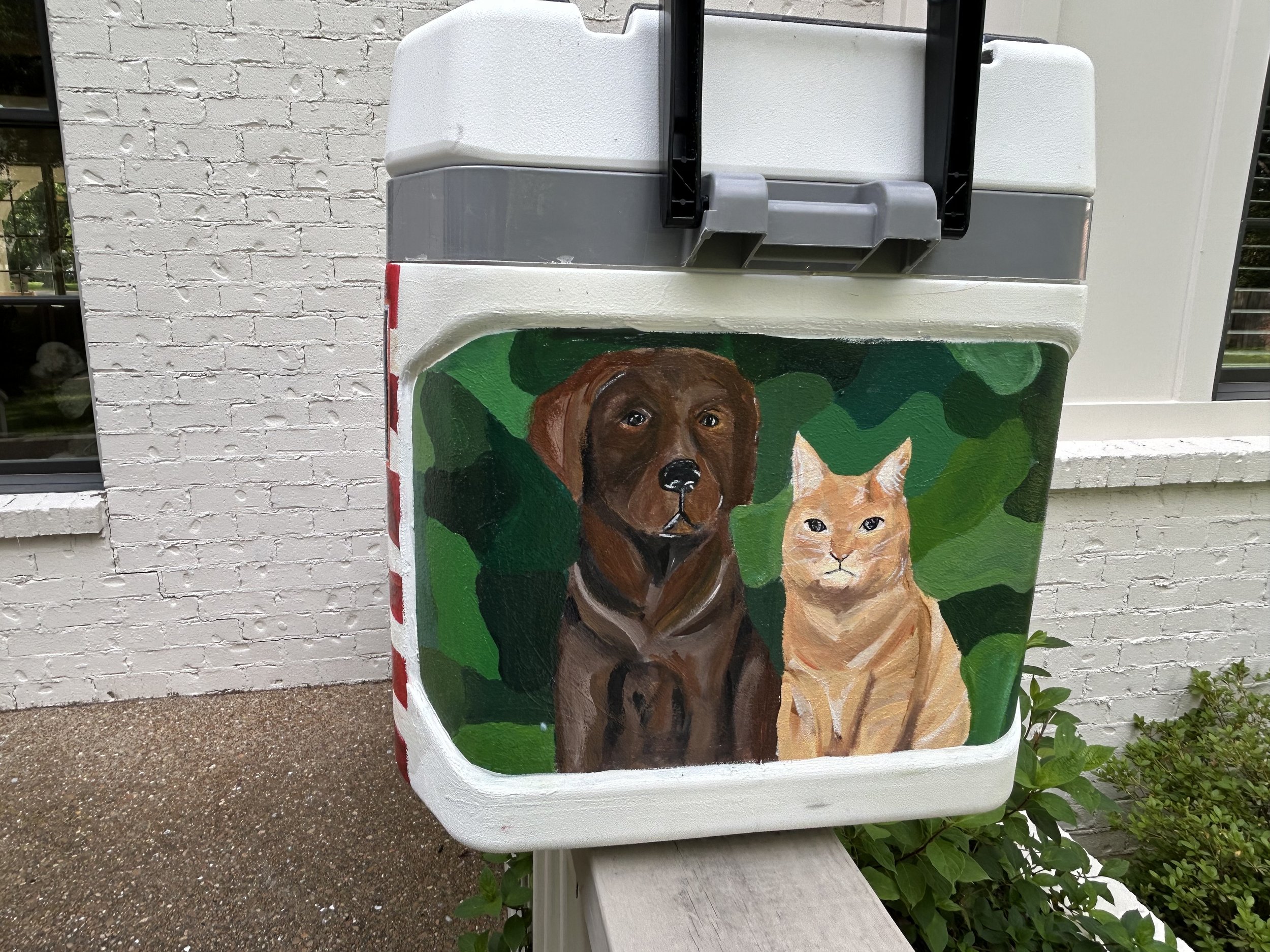 A painted mural of a brown dog and an orange cat on a white utility box in front of a brick wall with windows.