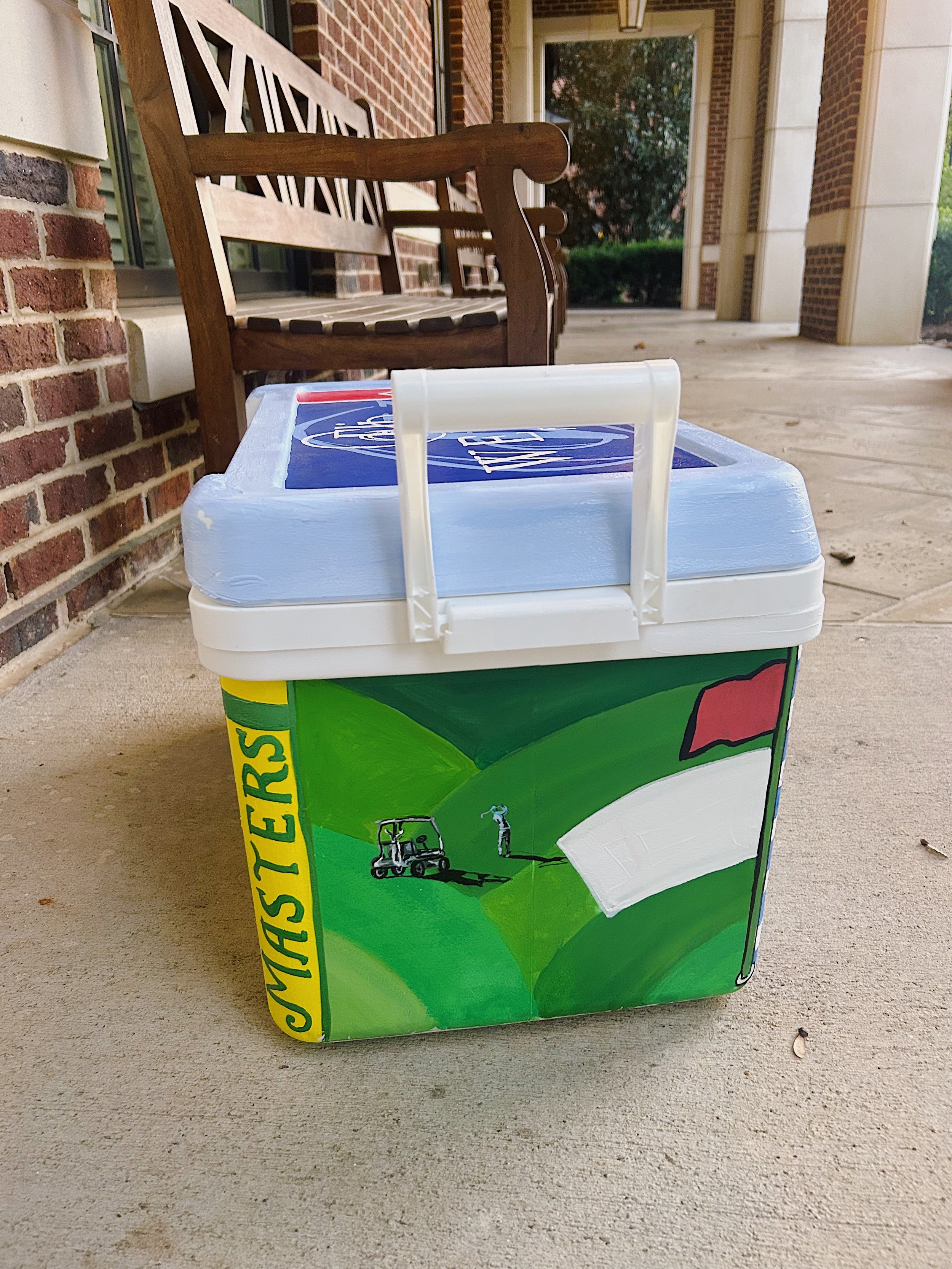 A painted styrofoam cooler box on a concrete porch, decorated with a green landscape scene, golf course imagery, and the word 'Masters' on the side.