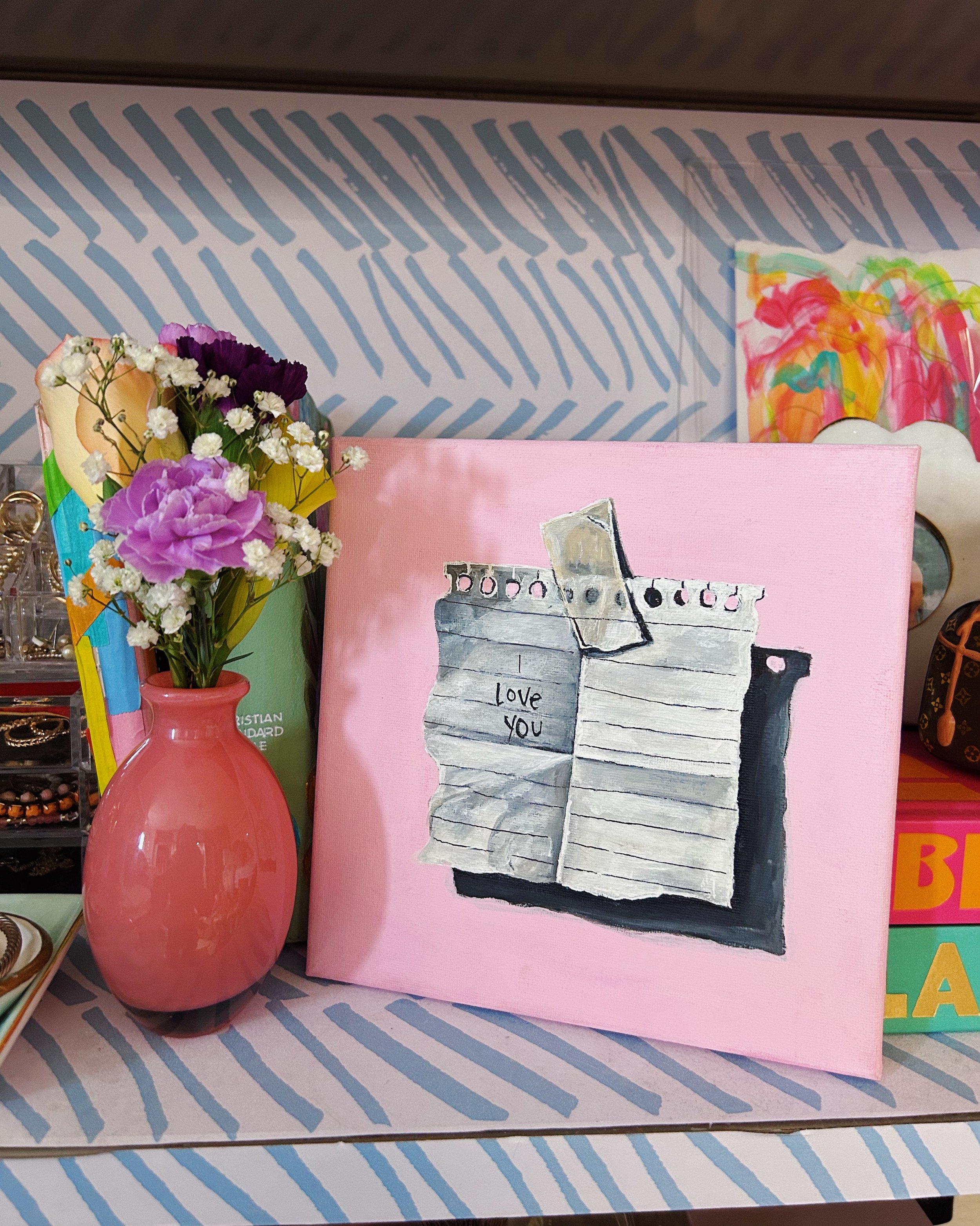 A pink-painted canvas with a drawing of a paper bag containing a phone, with the words "Love you" written on the bag. To the left, a pink vase holding a bouquet of flowers, including purple, white, and yellow blooms, sits on a striped surface.