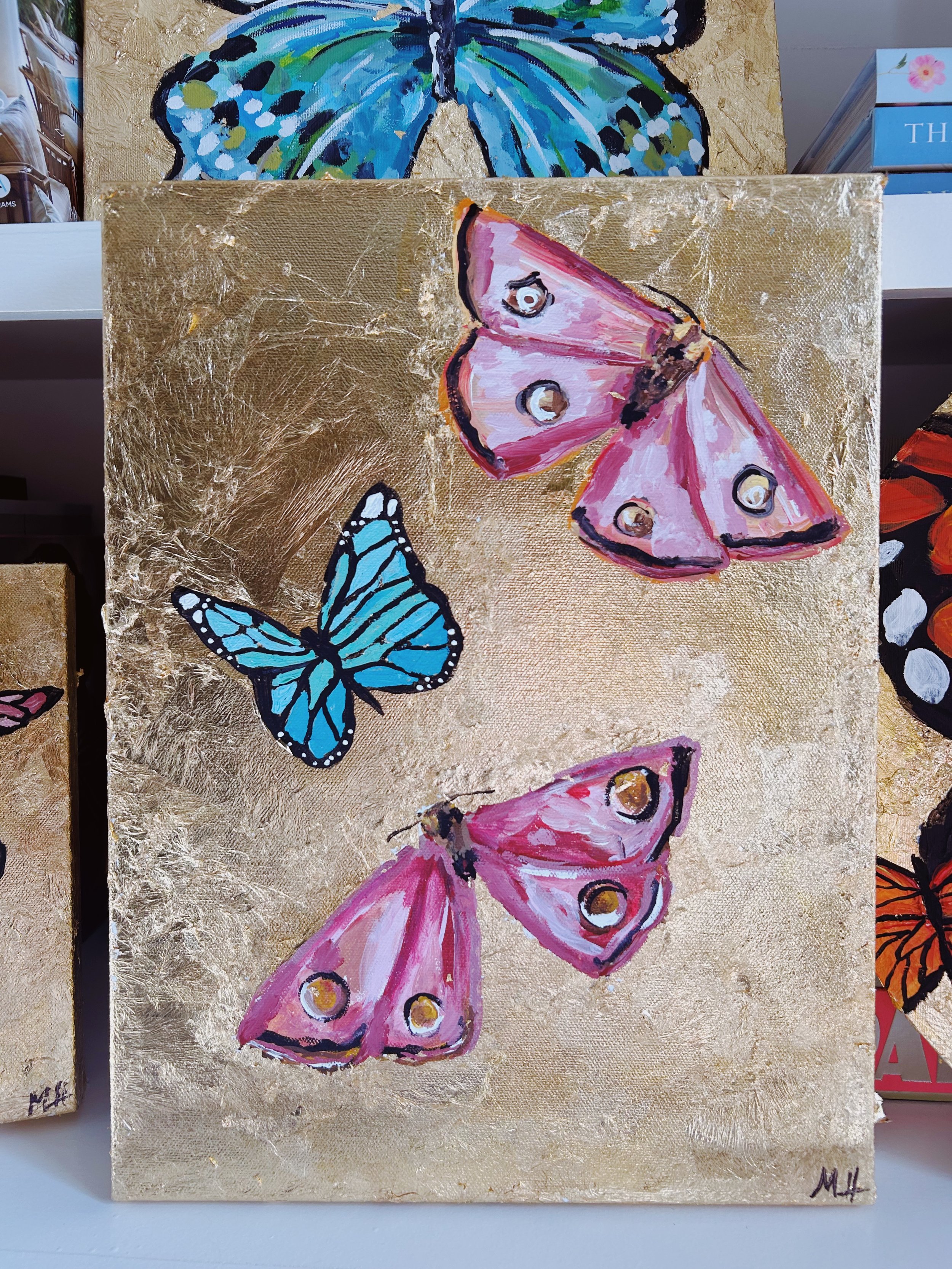 A painting of three butterflies on a gold background, with one butterfly in blue and two in pink, with a textured appearance.