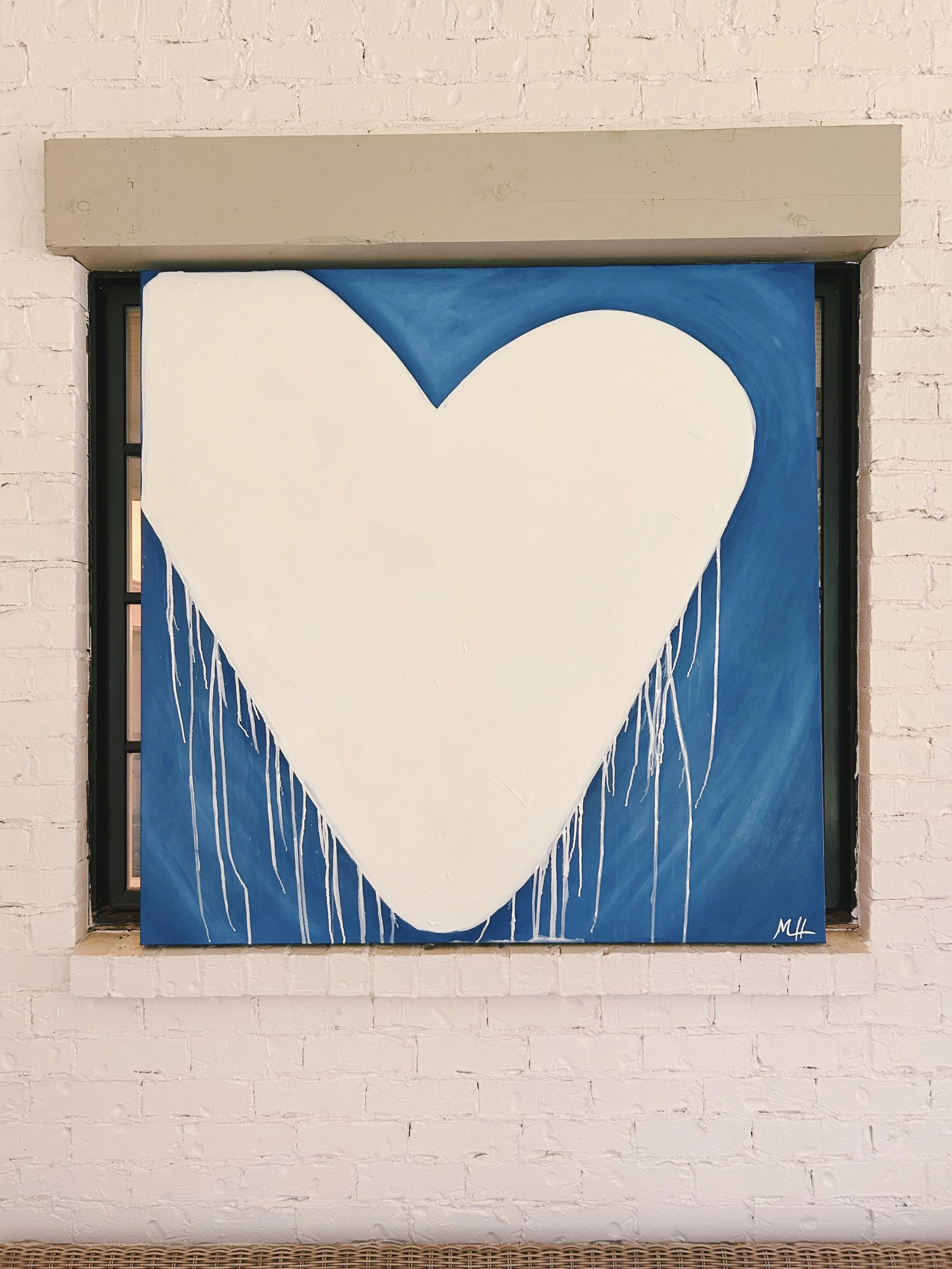 A painting of a large white heart with drips at the bottom, set against a blue background, mounted on a white brick wall.