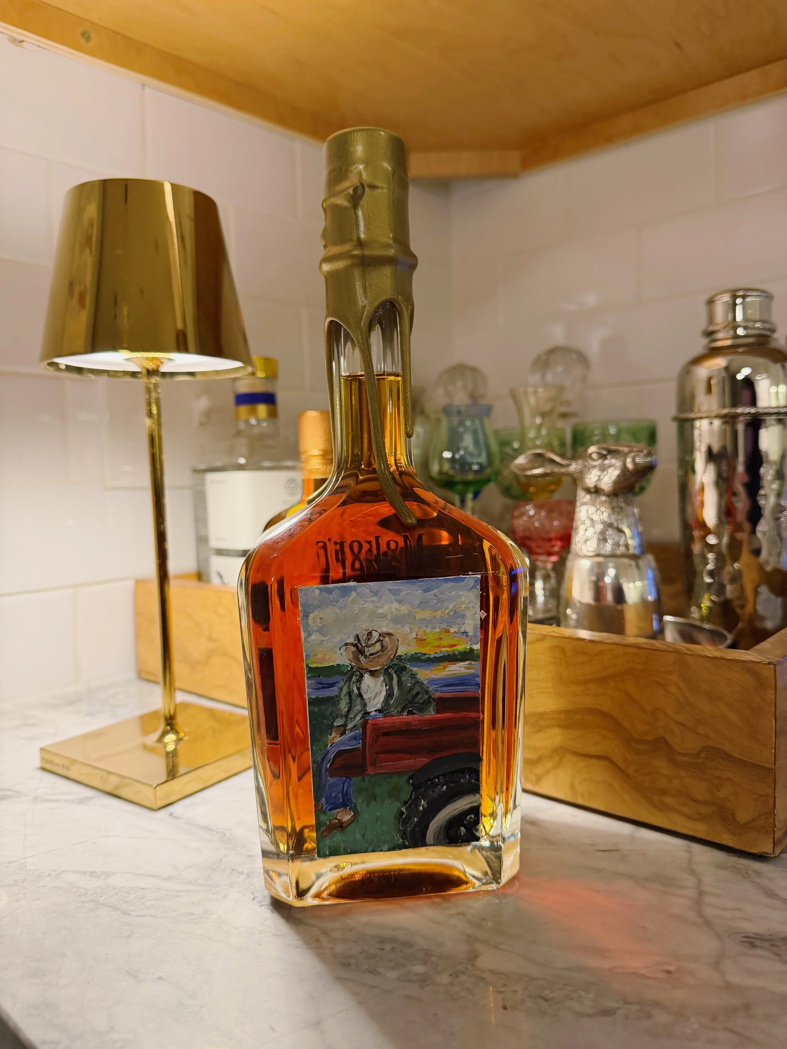 A glass bottle with orange liquid and a painted label featuring a person in a cowboy hat, sitting on a red wagon or cart, with a sunset or sunrise in the background. The bottle is placed on a kitchen countertop with various decorative items in the ba