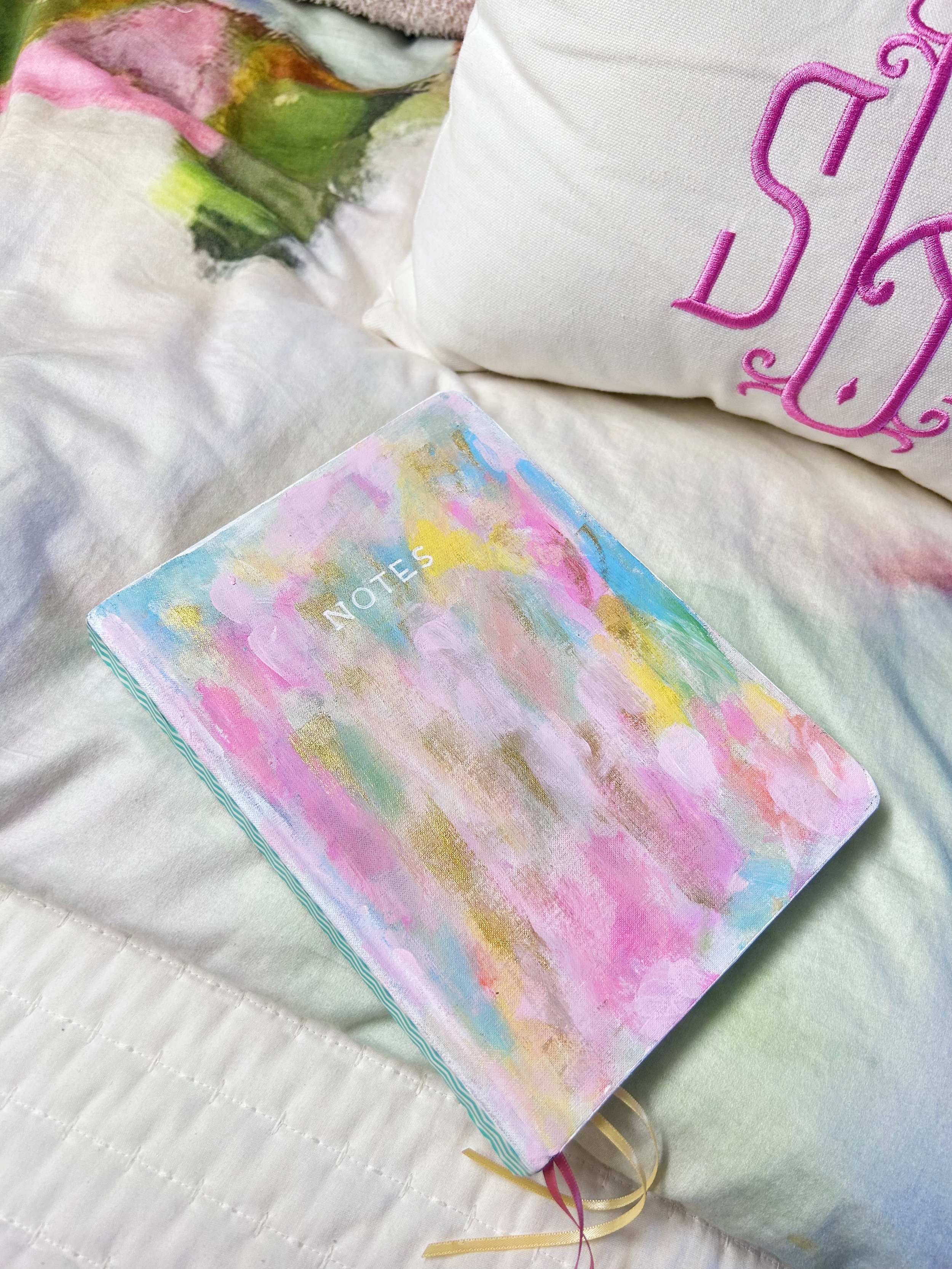 A pastel-colored notebook with 'NOTES' written on the cover, resting on a light-colored, textured bedspread. In the background, a white pillow with pink embroidery.