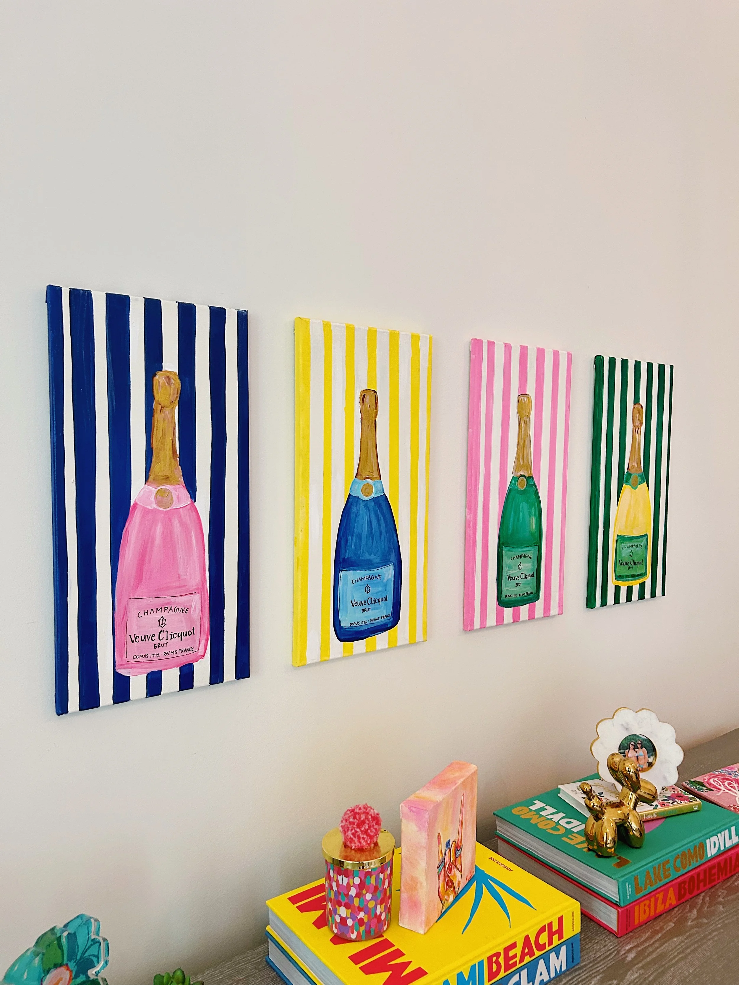 Four colorful paintings of champagne bottles with striped backgrounds, each in a different color scheme, hanging on a white wall.