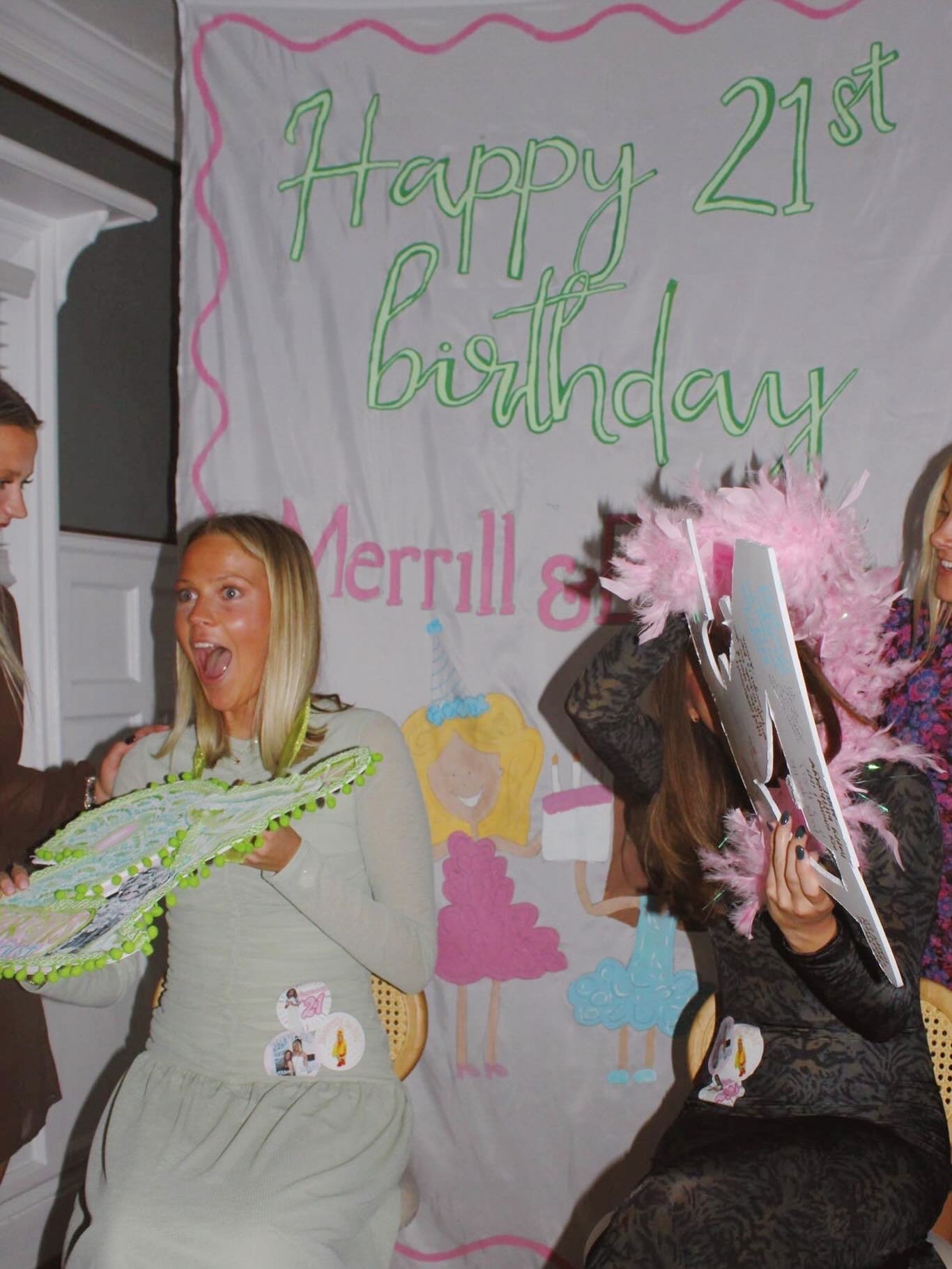 A group of women celebrating a 21st birthday, holding decorations and wearing party hats, standing in front of a colorful birthday banner that says 'Happy 21st birthday Merrill &' with cartoon images of women and birthday cakes.
