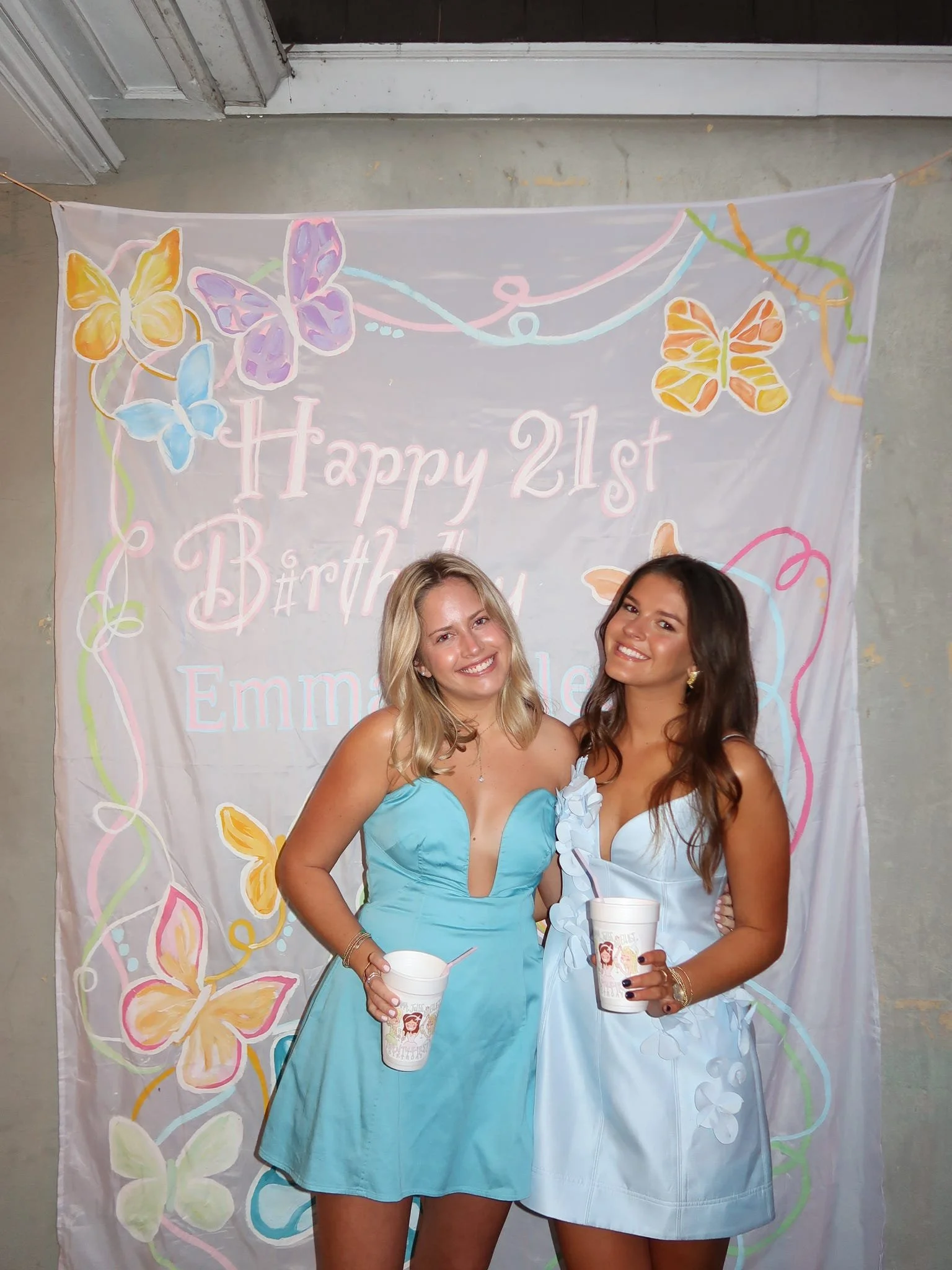 Two women celebrating at a birthday party, standing in front of a decorated backdrop with butterflies and the text 'Happy 21st Birthday' and the name 'Emma'. They are holding drinks and smiling.