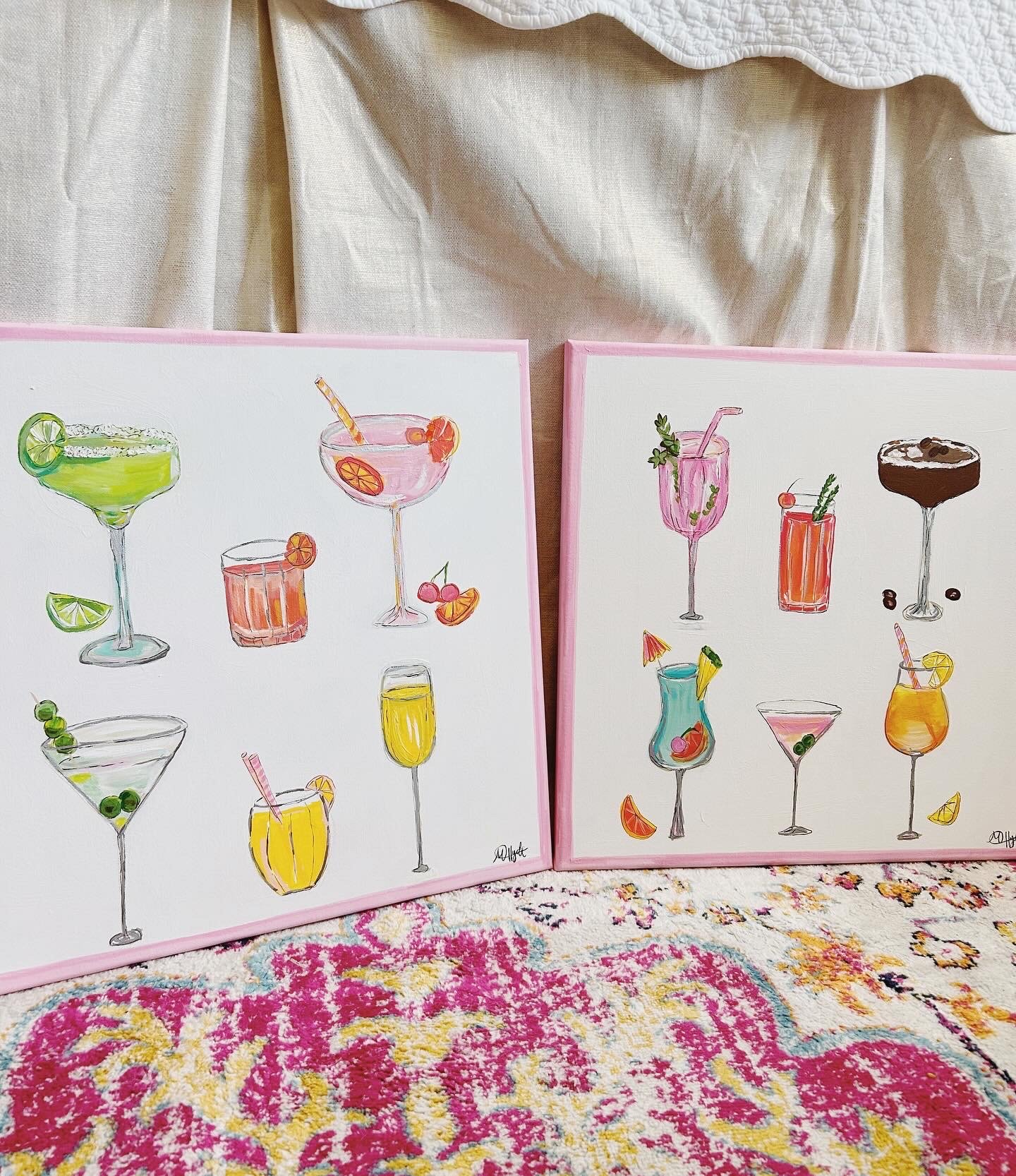 Two framed watercolor paintings of various colorful cocktails, placed on a pink surface with a decorative colorful rug underneath and light-colored curtains behind.