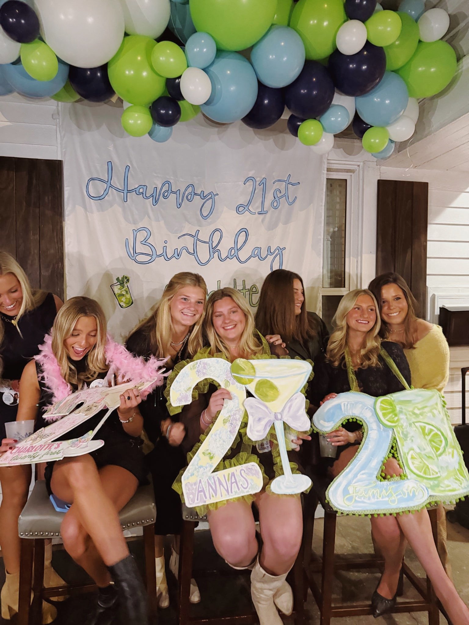 Group of women celebrating 21st birthday with colorful balloons, signs, and a decorated backdrop reading 'Happy 21st Birthday'.
