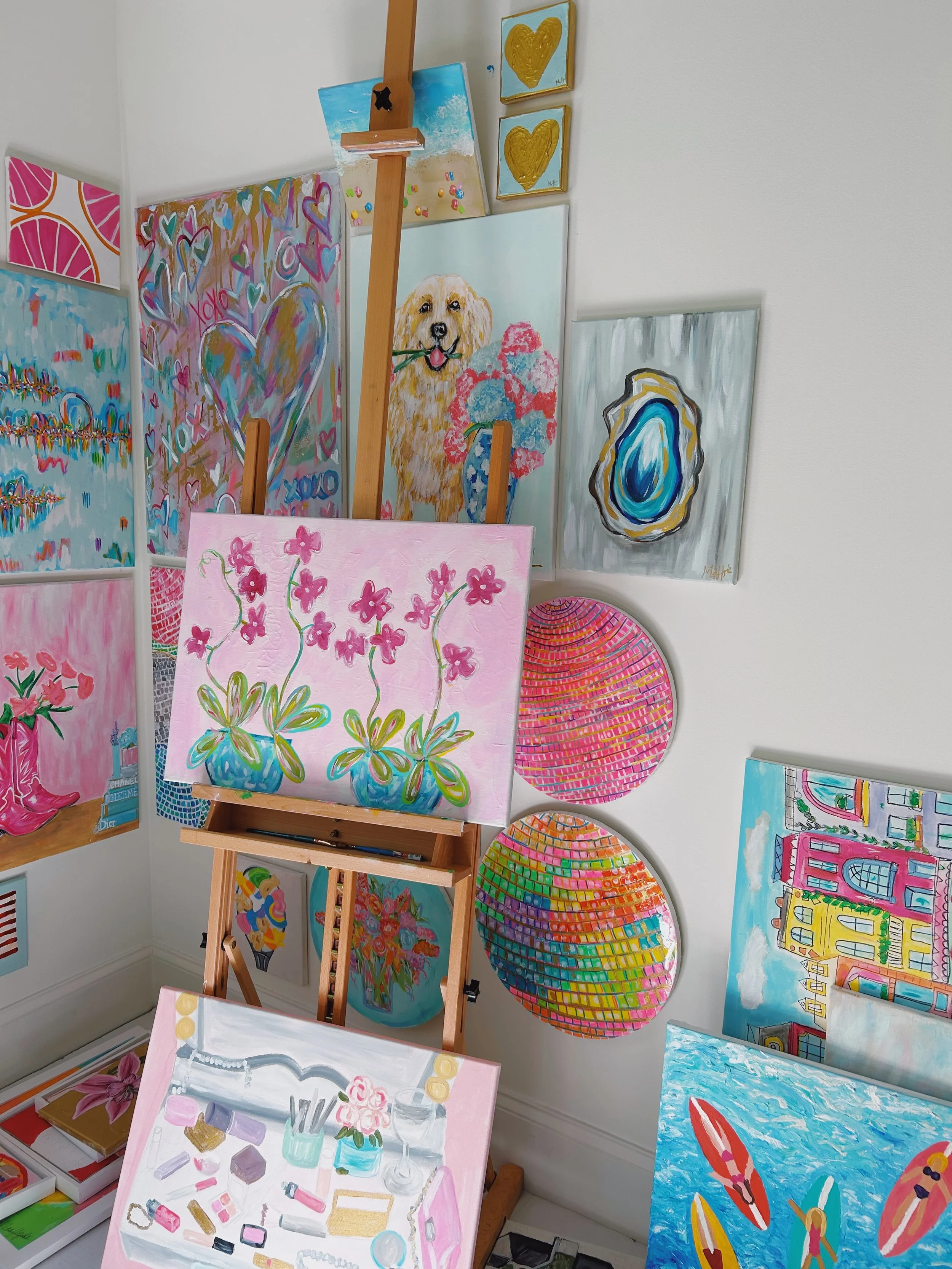 Collection of colorful paintings and artworks displayed on a white gallery wall and easel, including abstract, floral, animal, and cityscape themes.