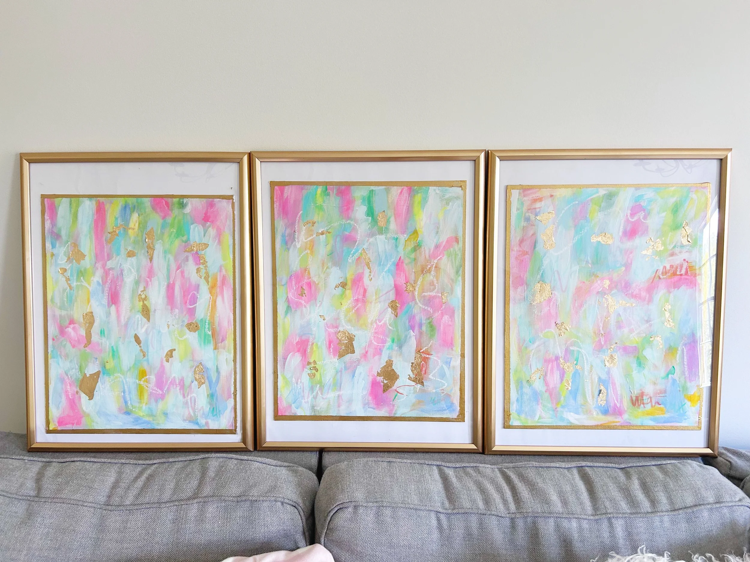 Three colorful abstract paintings with gold leaf accents in gold frames, leaning against a plain wall on a gray couch.