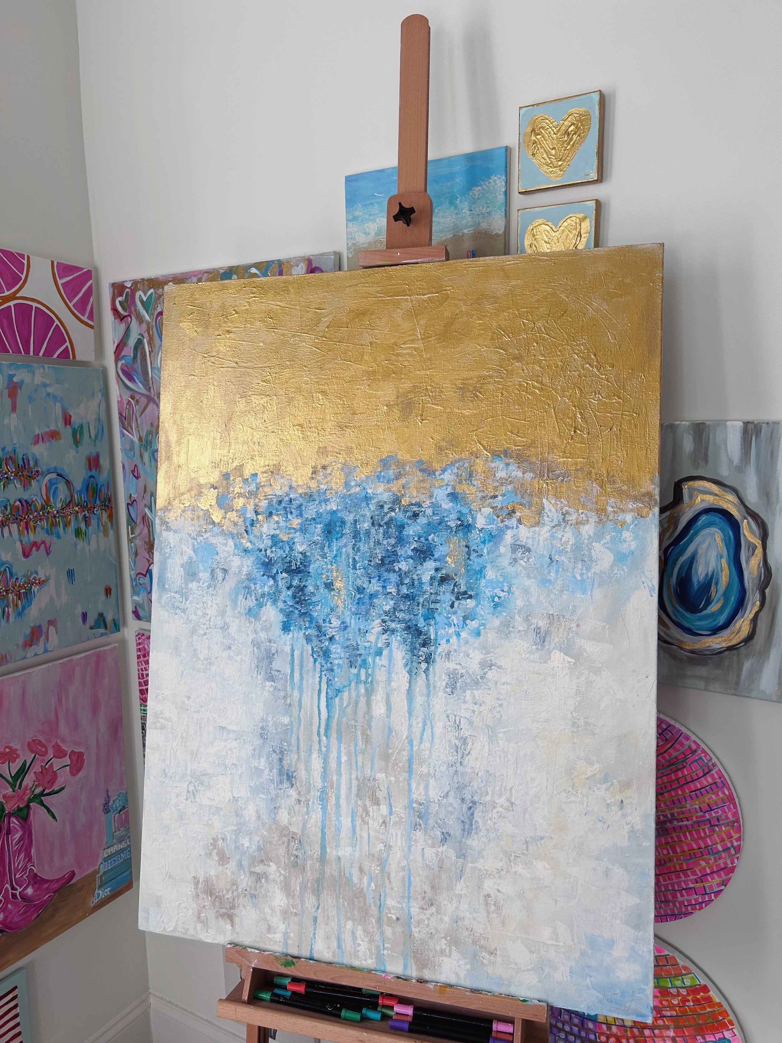 An abstract painting on an easel with a gold top and a blue and white bottom, dripping paint, in a room with other colorful paintings and wall decorations.