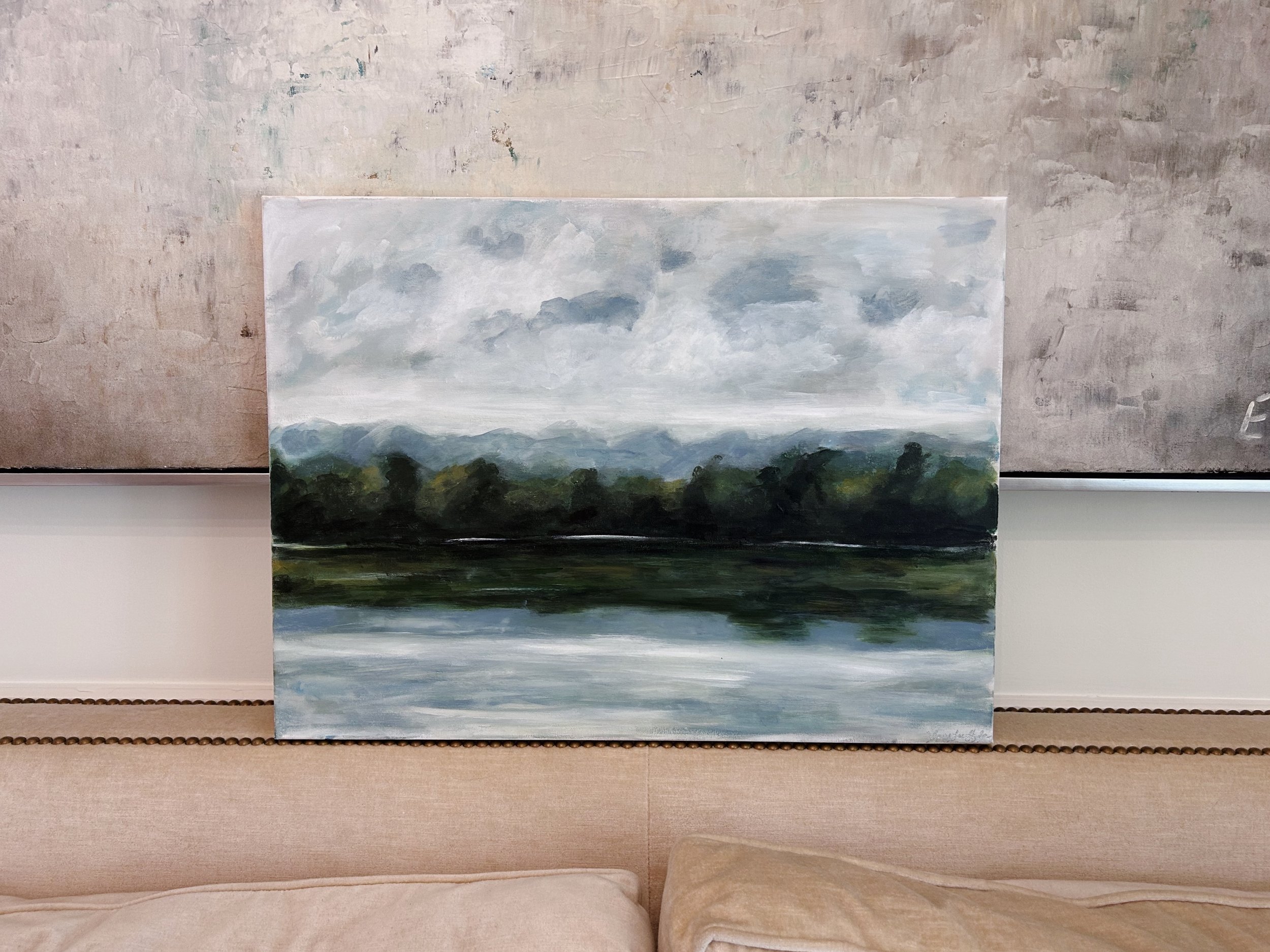 A landscape painting of a lake with trees and mountains in the background, under a cloudy sky.