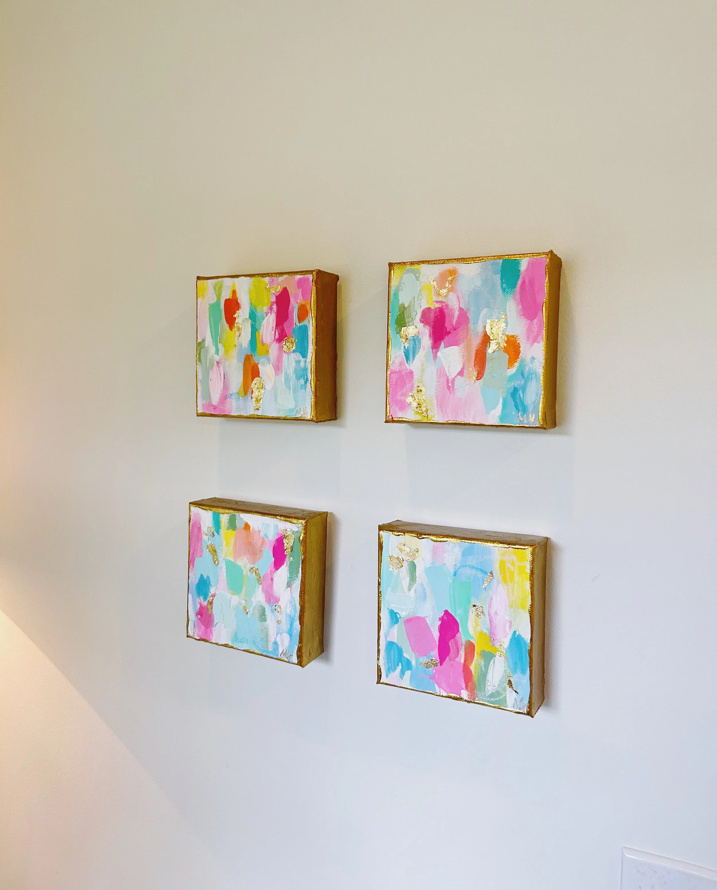 Four small colorful abstract paintings with gold accents arranged in a 2x2 grid on a white wall.