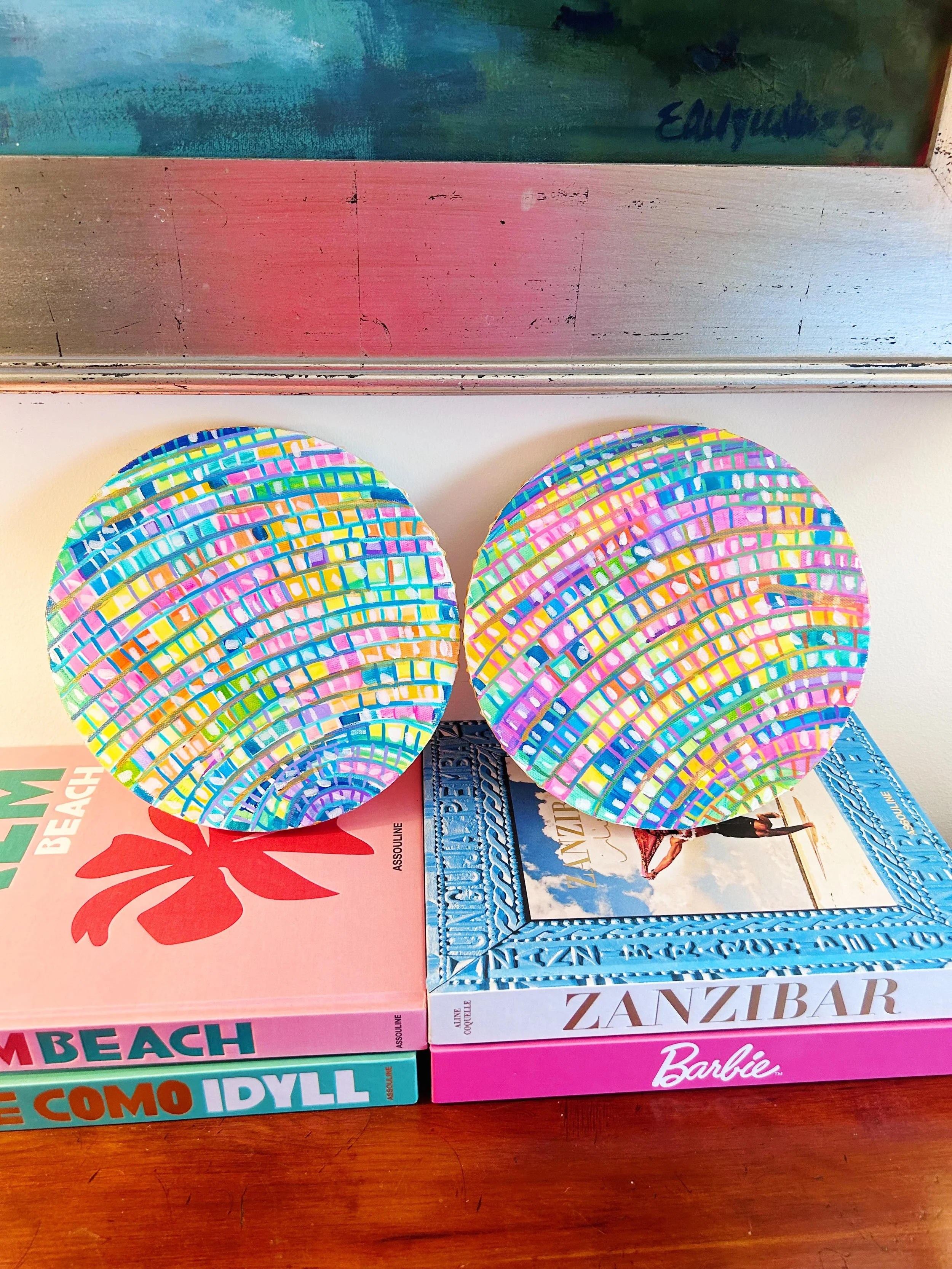 Two round, colorful mosaic-style decorative objects placed on top of framed books about Zanzibar, beach, and Italy, on a wooden surface beneath a reflective metallic surface.
