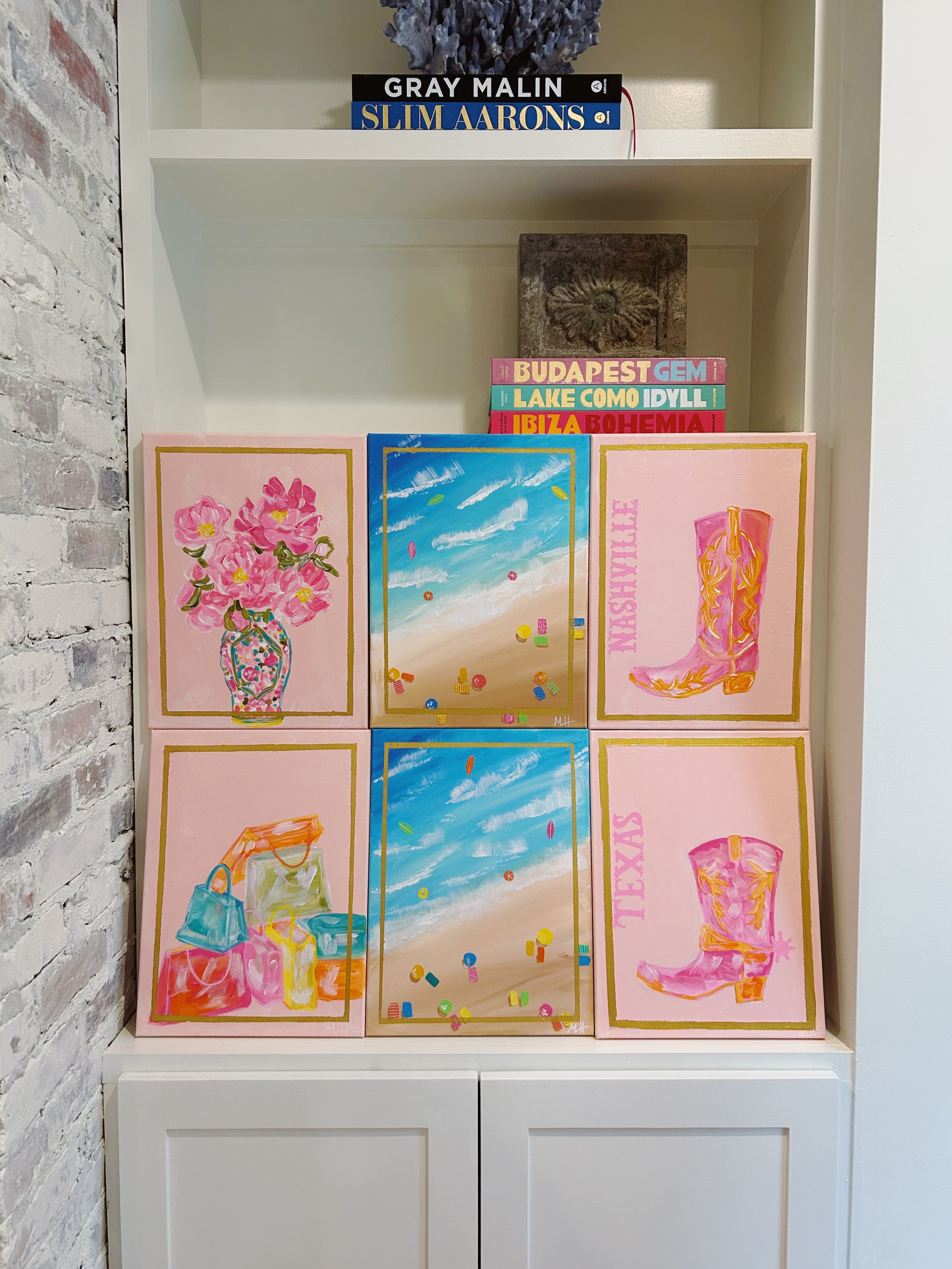 Six colorful paintings arranged in a grid on a white cabinet, with a bookshelf above holding travel books and decorative items.