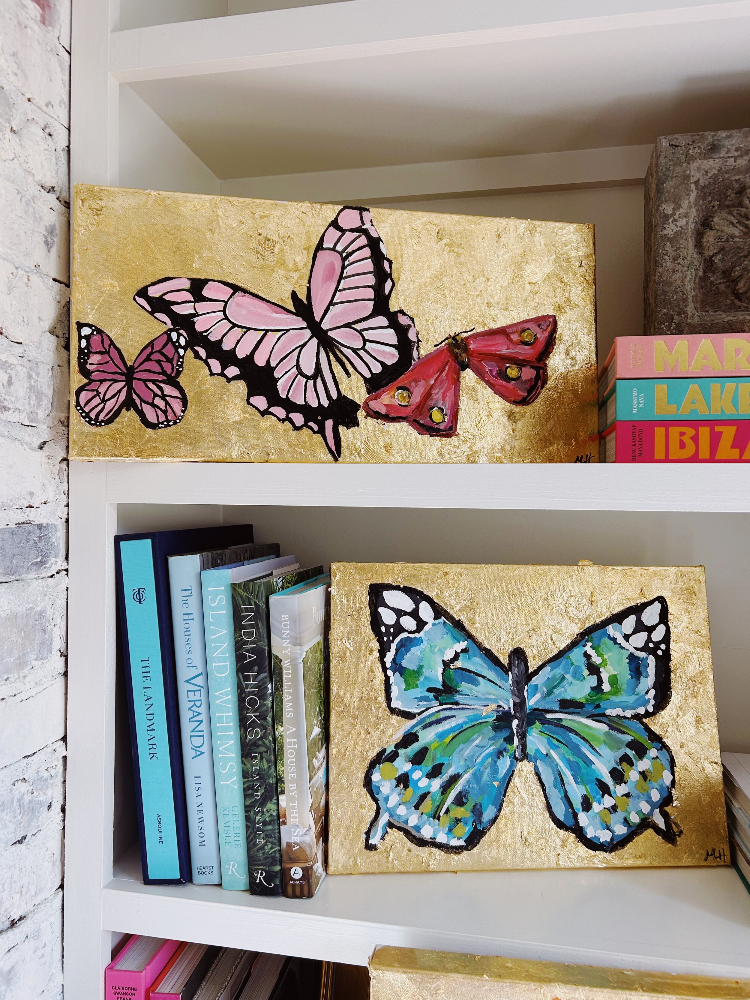 Two decorative paintings of butterflies on a white bookshelf, with books around and behind them. The top butterfly is pink with black outlines, and the bottom butterfly is blue and green with black outlines, both on gold backgrounds.