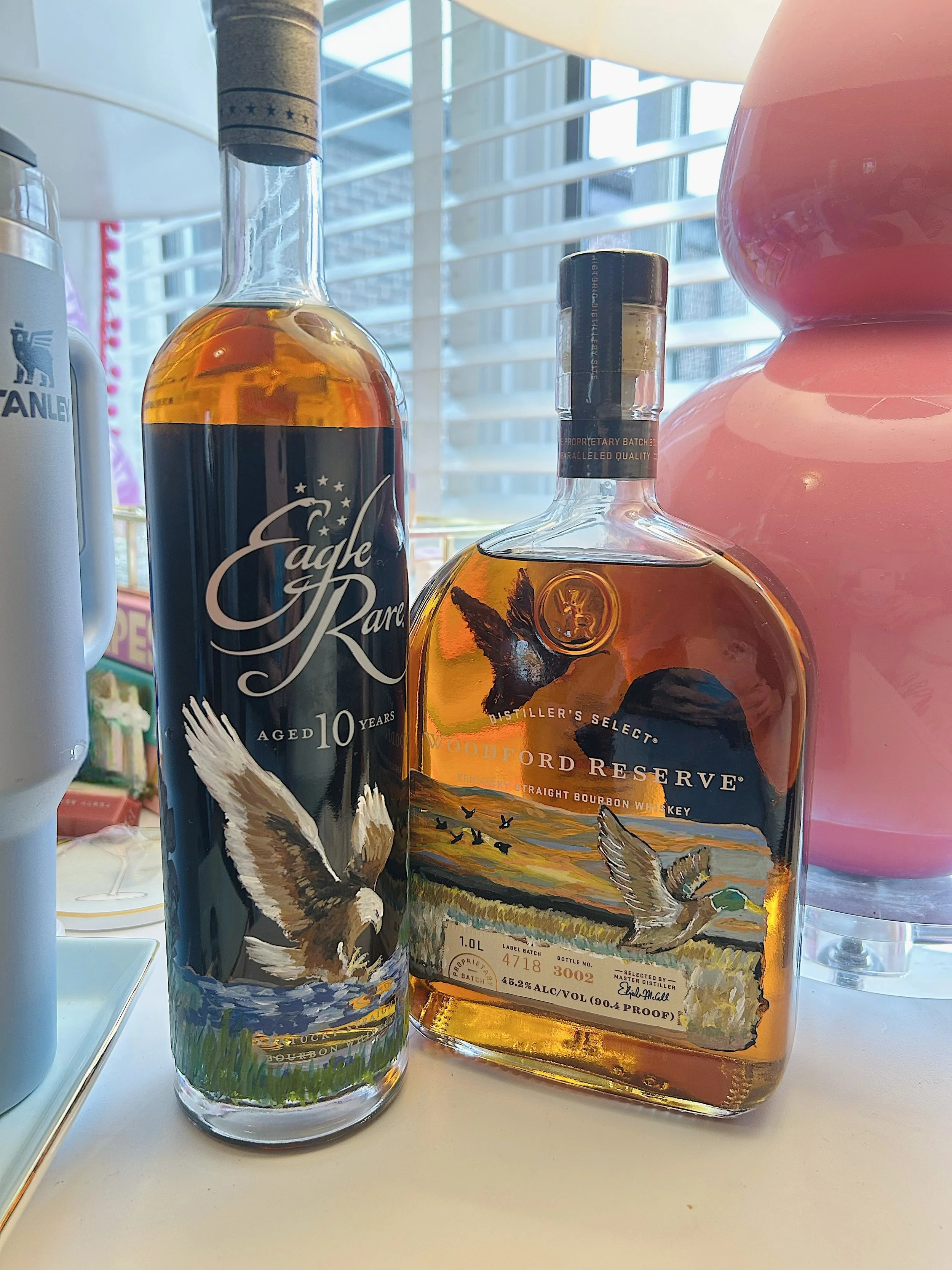 Two bottles of whiskey, one Eagle Rare aged 10 years and one Woodford Reserve Kentucky straight bourbon, sitting on a white surface near a window with blinds.