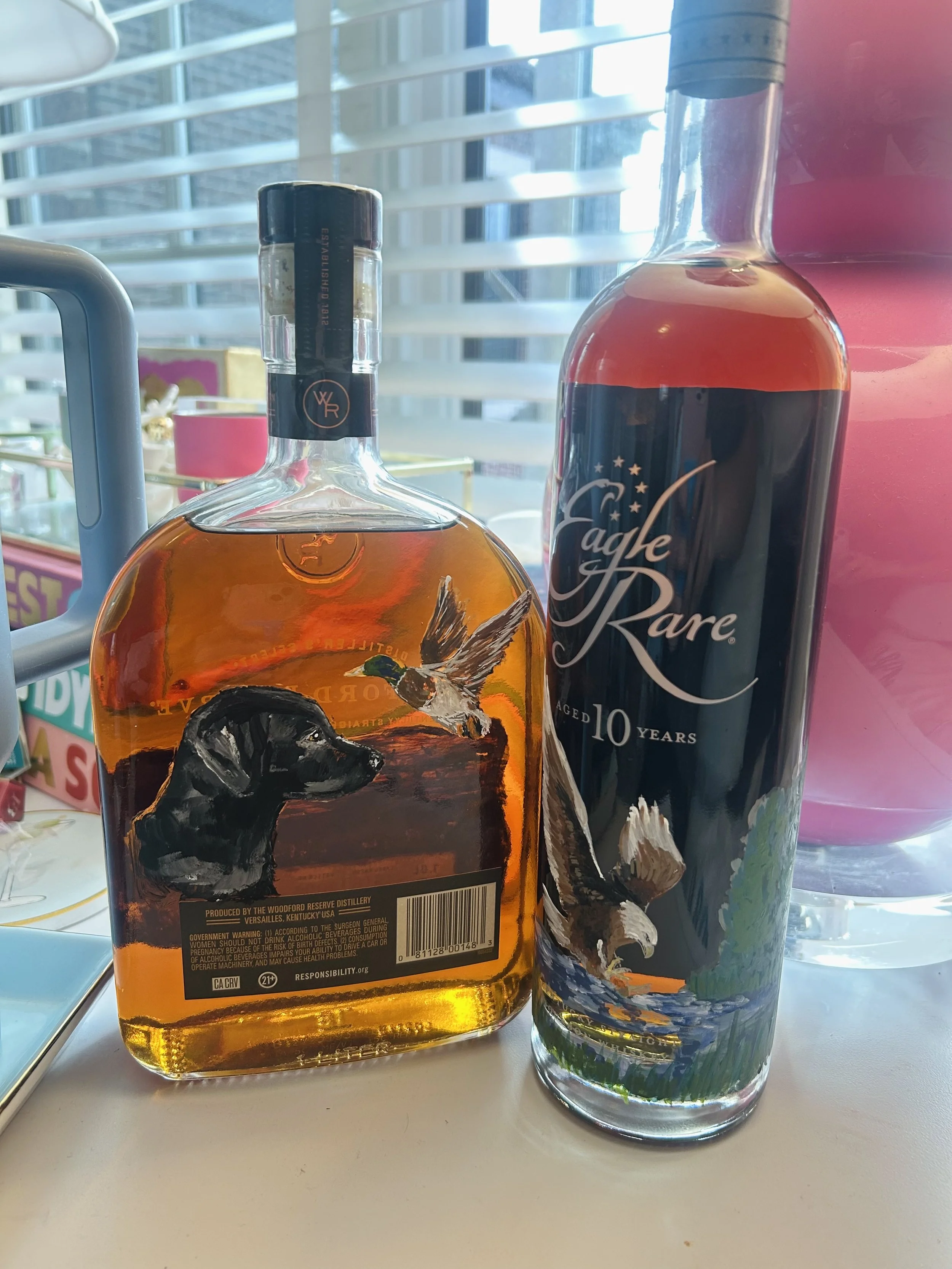 Two bottles of alcohol on a window sill. The left bottle has a painted dog and a bird illustration, the right bottle has 