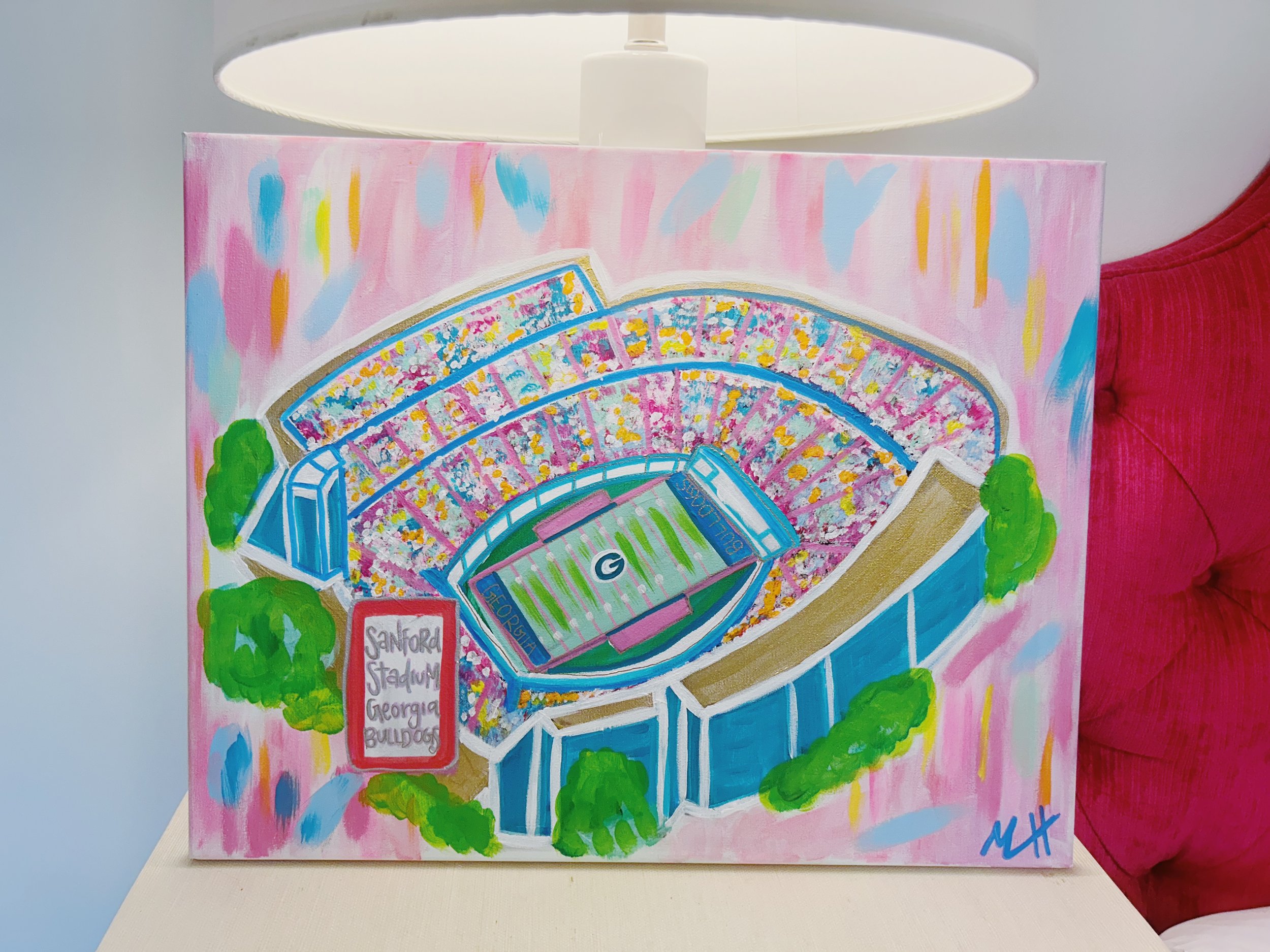 A colorful painting of Sanford Stadium in Georgia with green trees around it, showing the field with a large G logo in the center and the stands filled with yellow, red, purple, and blue seats, against a pink and pastel background.
