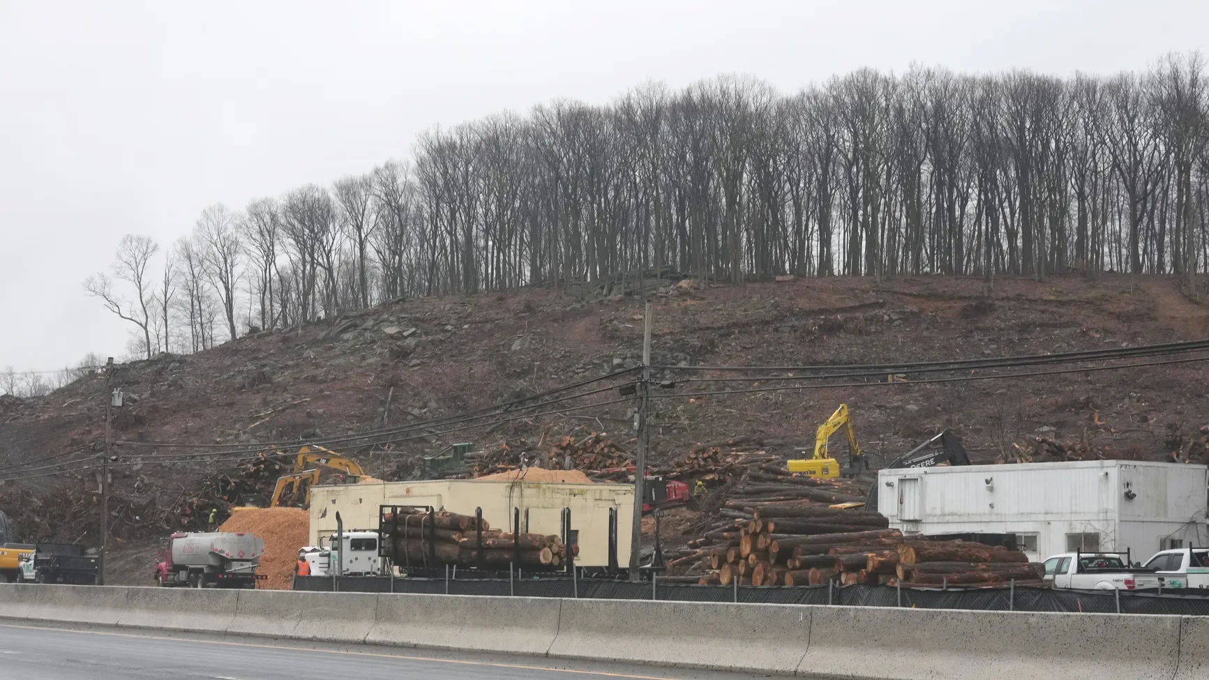 Construction vehicles and logs near a hillside with leafless trees, on a cloudy day.