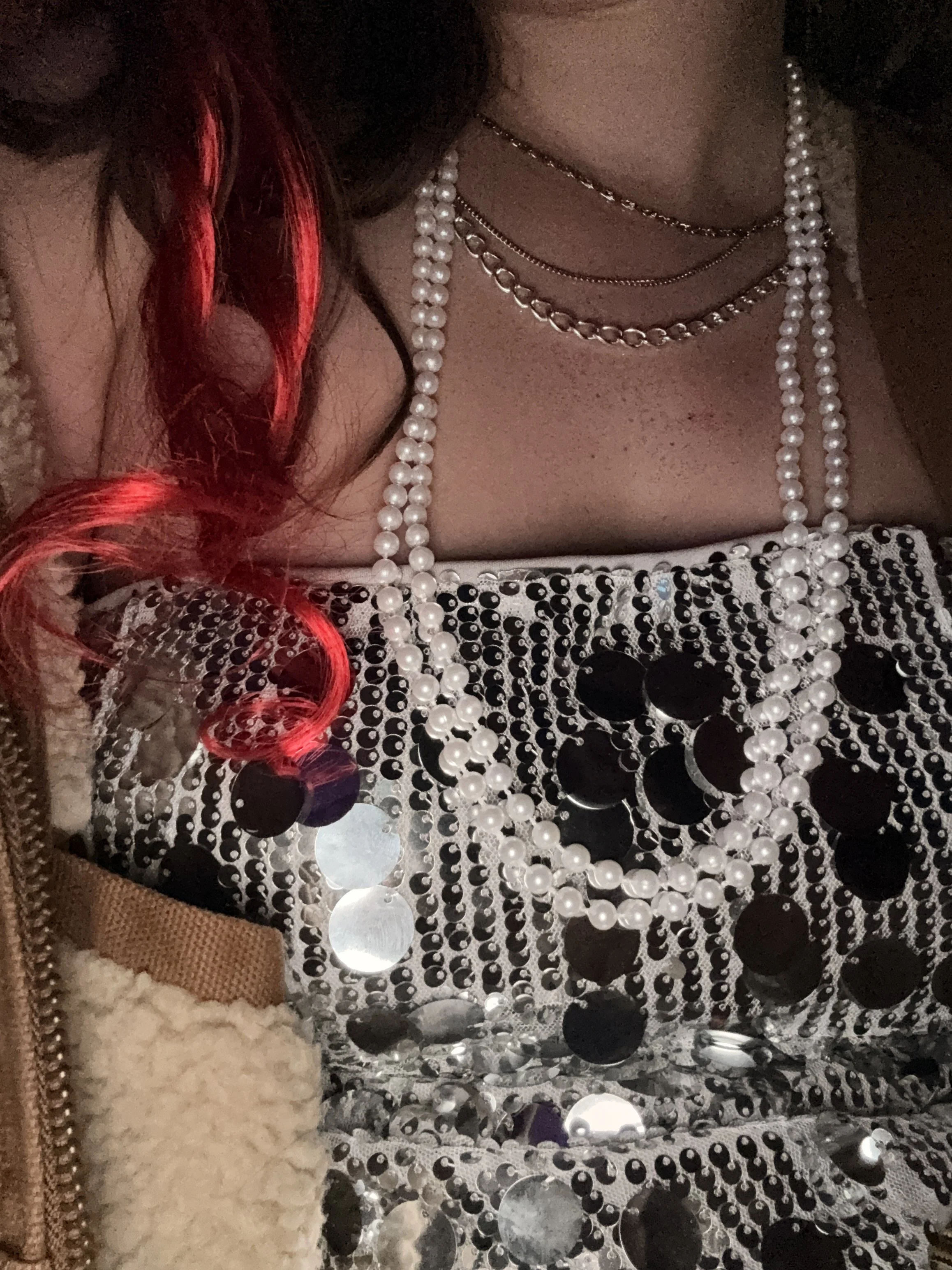Close-up of a woman's chest and shoulder with layered necklaces, a sequined dress with black dots, and a beige coat with fur lining, with red hair curls visible.