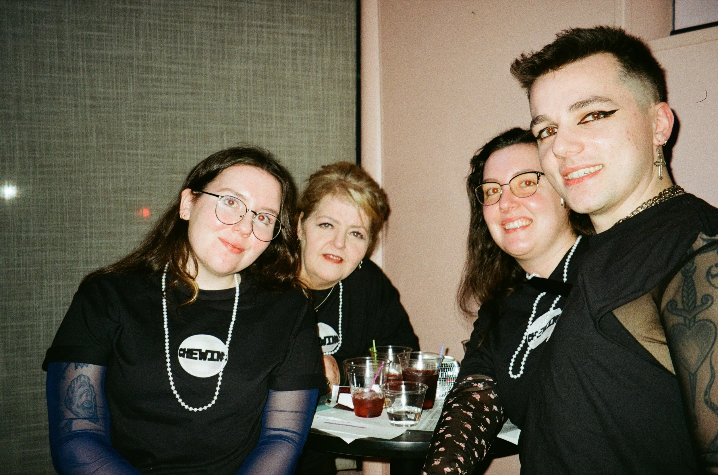 Four people sitting together at a table during a social gathering, smiling at the camera. They are wearing black shirts with a white logo, necklaces, and some have glasses and tattoos. There are drinks on the table.