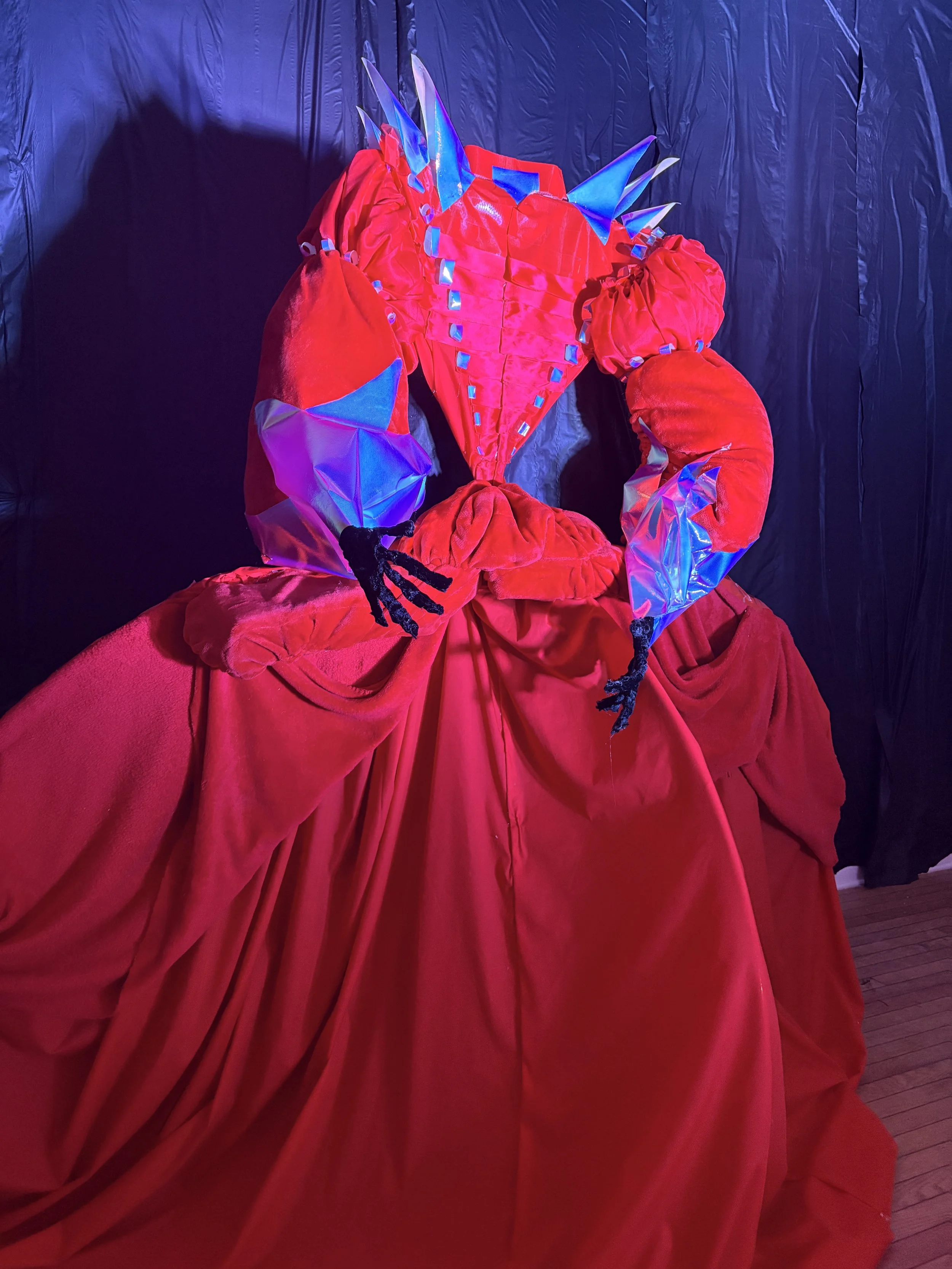 A costume of a lady in a red dress with a large, voluminous skirt, featuring a corset-style bodice with shimmering embellishments, puffy sleeves, and metallic blue accents on the sleeves, displayed against a dark backdrop.