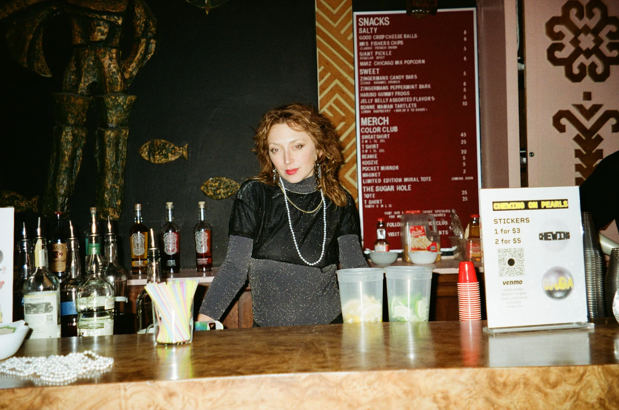 A woman with curly red hair wearing a black shimmery top and layered pearl necklaces standing behind a bar counter. The bar has various bottles of liquor, a container of straws, and cups with lime wedges. In the background, there is a red menu board 