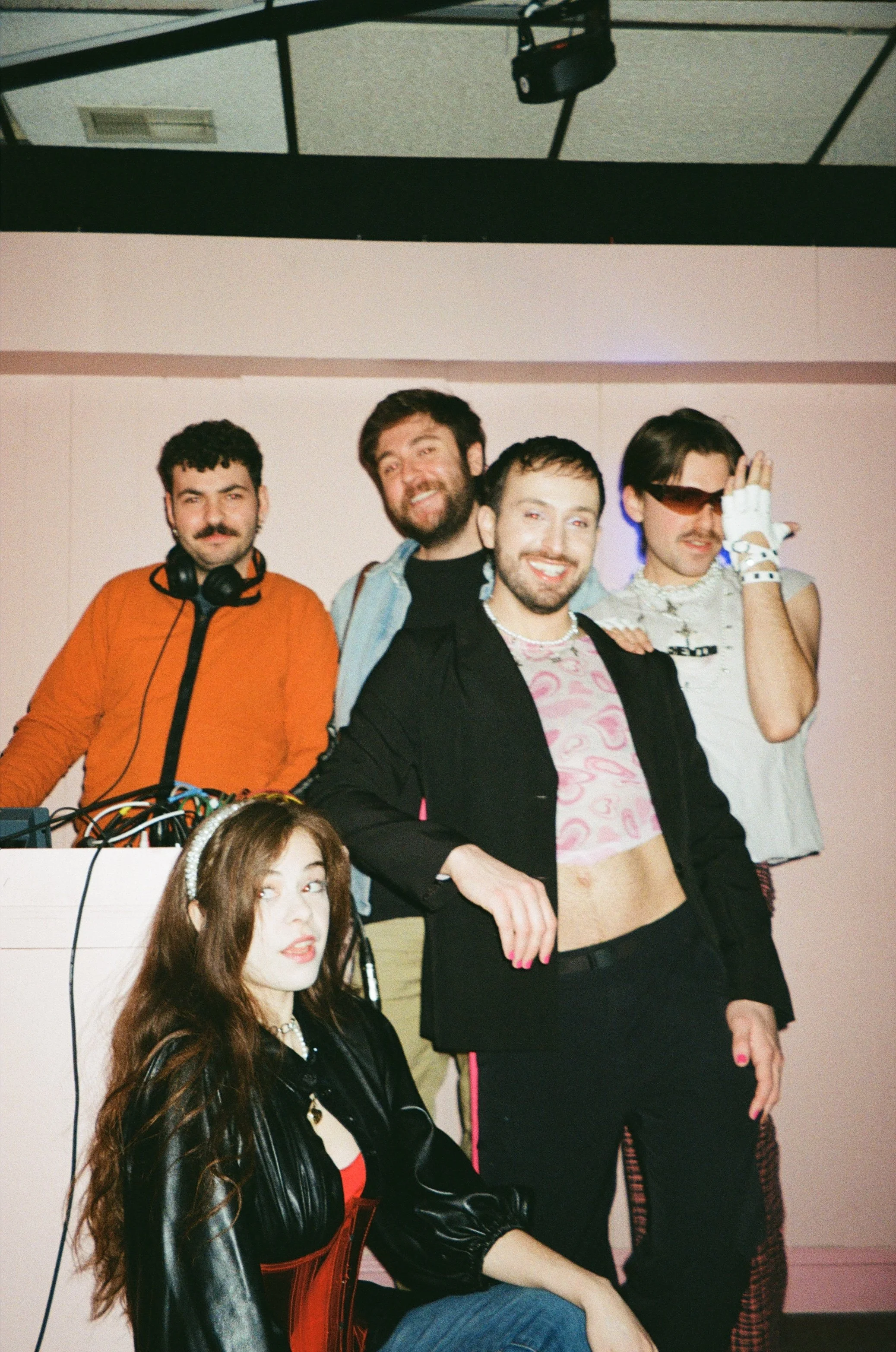 Group of five young adults posing together—two women and three men—at an indoor event, with one dressed in casual fashion, one in a leather jacket, and one wearing sunglasses and gloves.