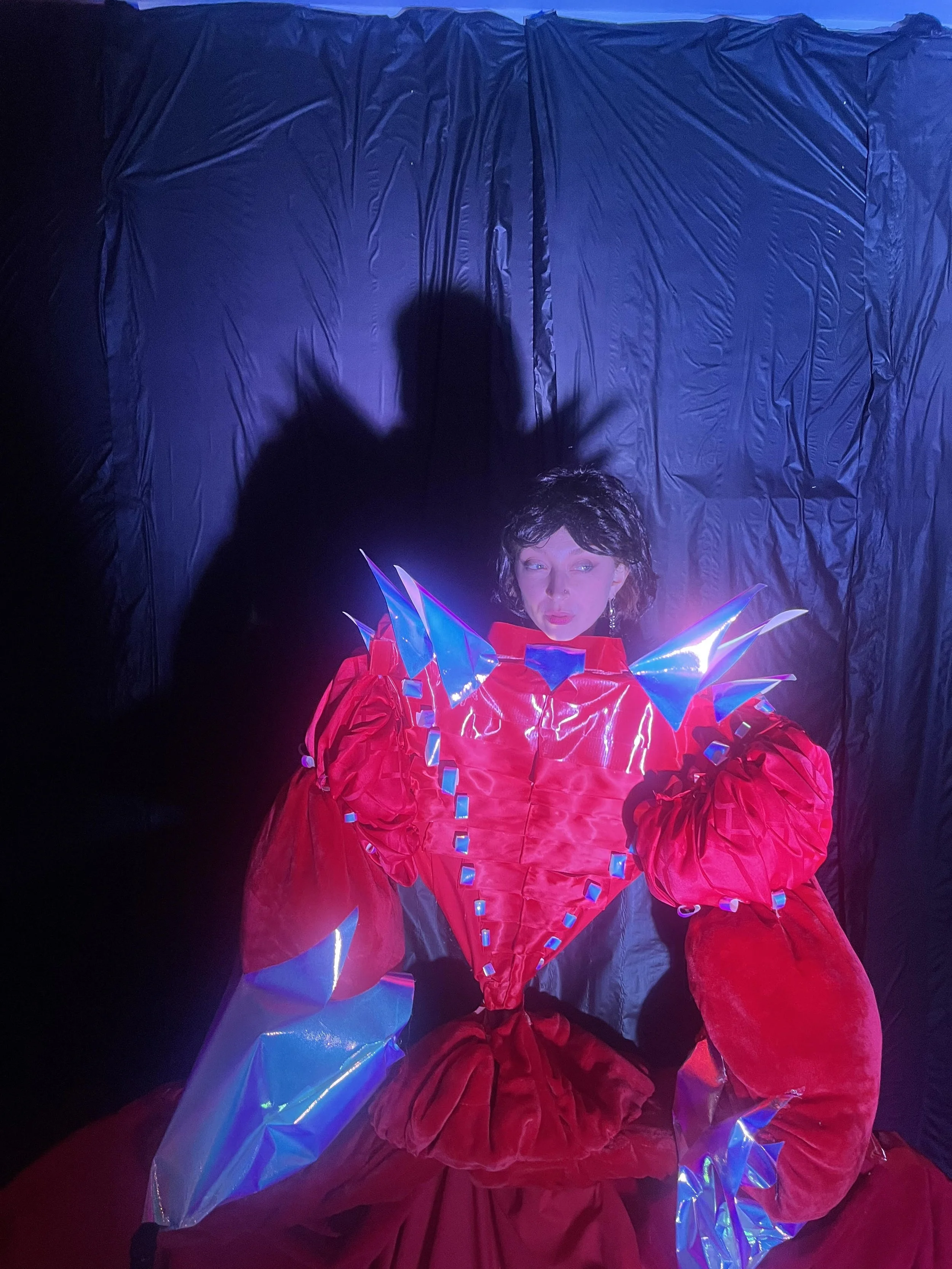 A woman dressed in a futuristic red costume with blue and transparent accents, standing against a dark background with a blue curtain, illuminated by colorful lighting.