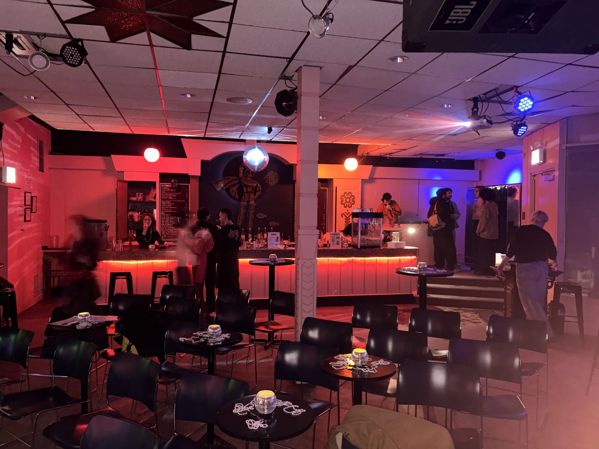 Interior of a bar or nightclub with red and blue lighting. Empty tables with black chairs are in the foreground, some with glasses and napkins. The bar counter is in the background with a few people standing around, and a DJ or bartender behind the c