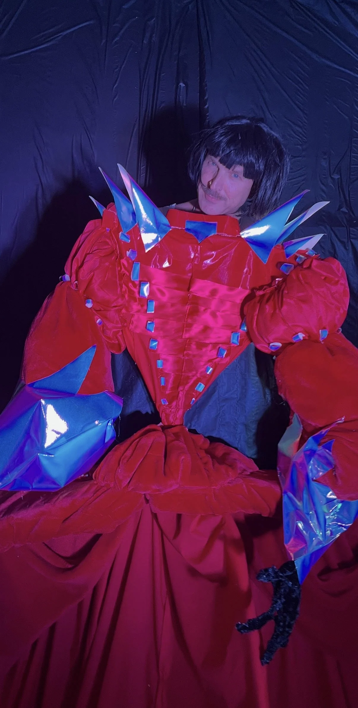 Person dressed in a vibrant red, costume with shiny blue accents and spikes, lying down on a black background, making a playful face.