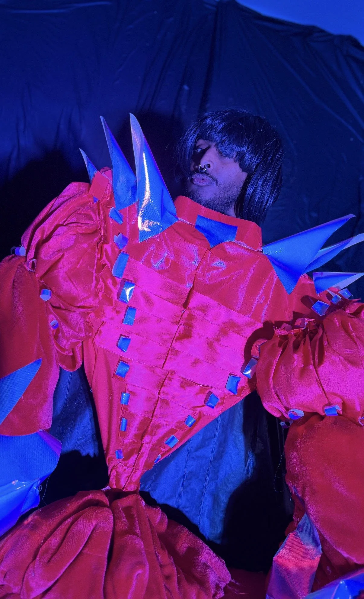 Person wearing a red, shiny, spiked costume with blue accents, posing against a dark background.