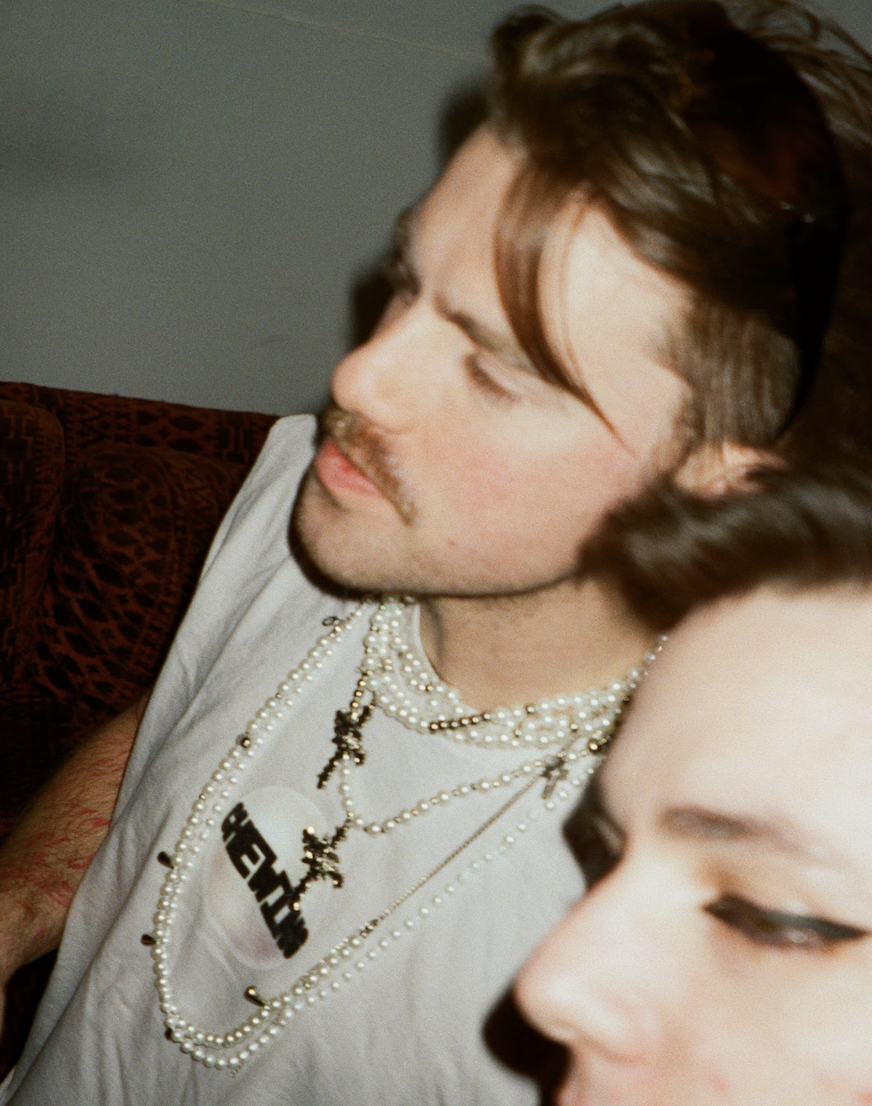 Close-up of a man with brown hair, a mustache, and layered pearl necklaces, sitting next to a woman with makeup, both with relaxed expressions.