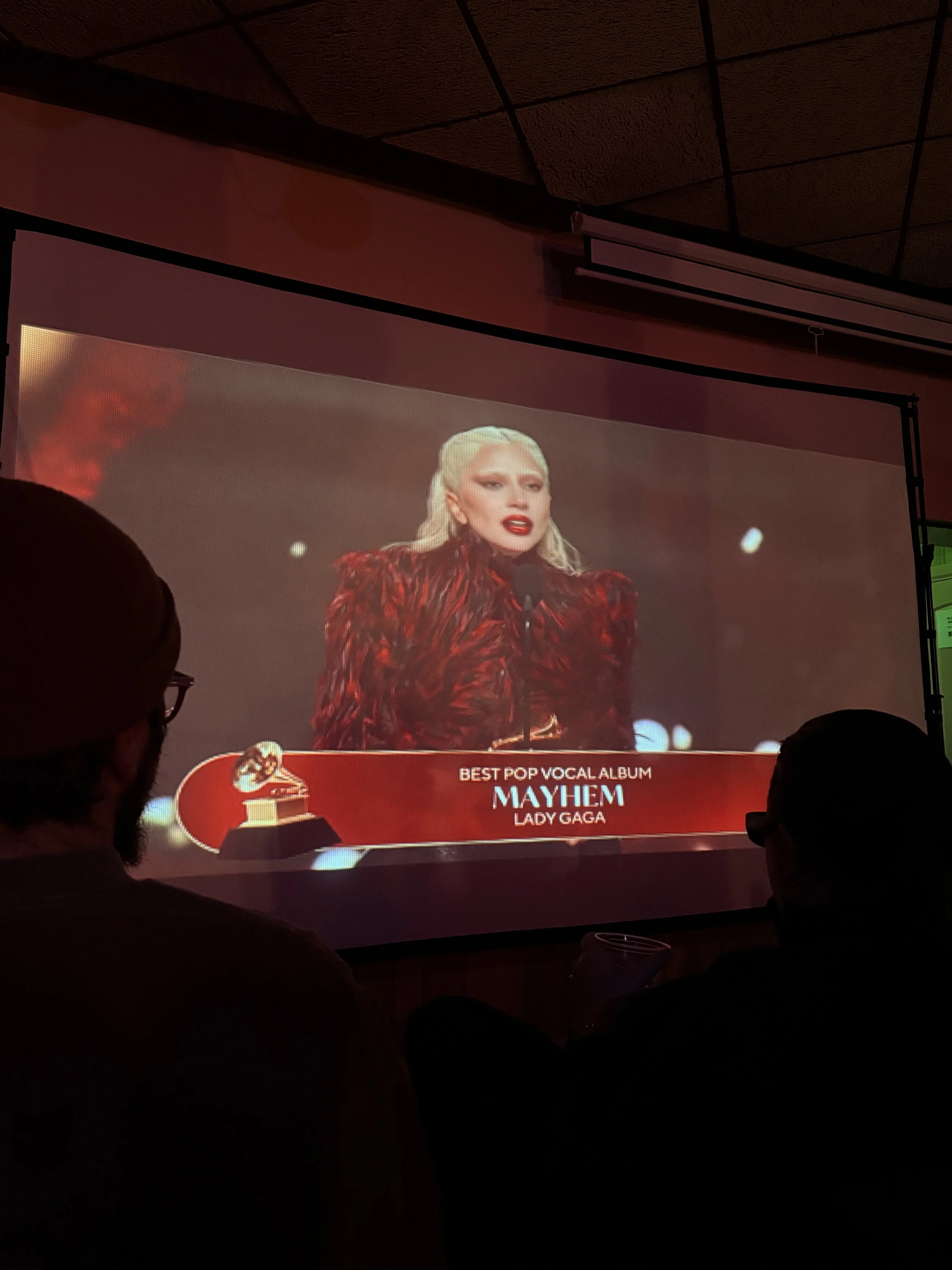 Image of a woman with blonde hair and red lipstick, wearing a red and black outfit, speaking at an award ceremony. The screen displays the text "Best Pop Vocal Album Lady Gaga Mayhem".