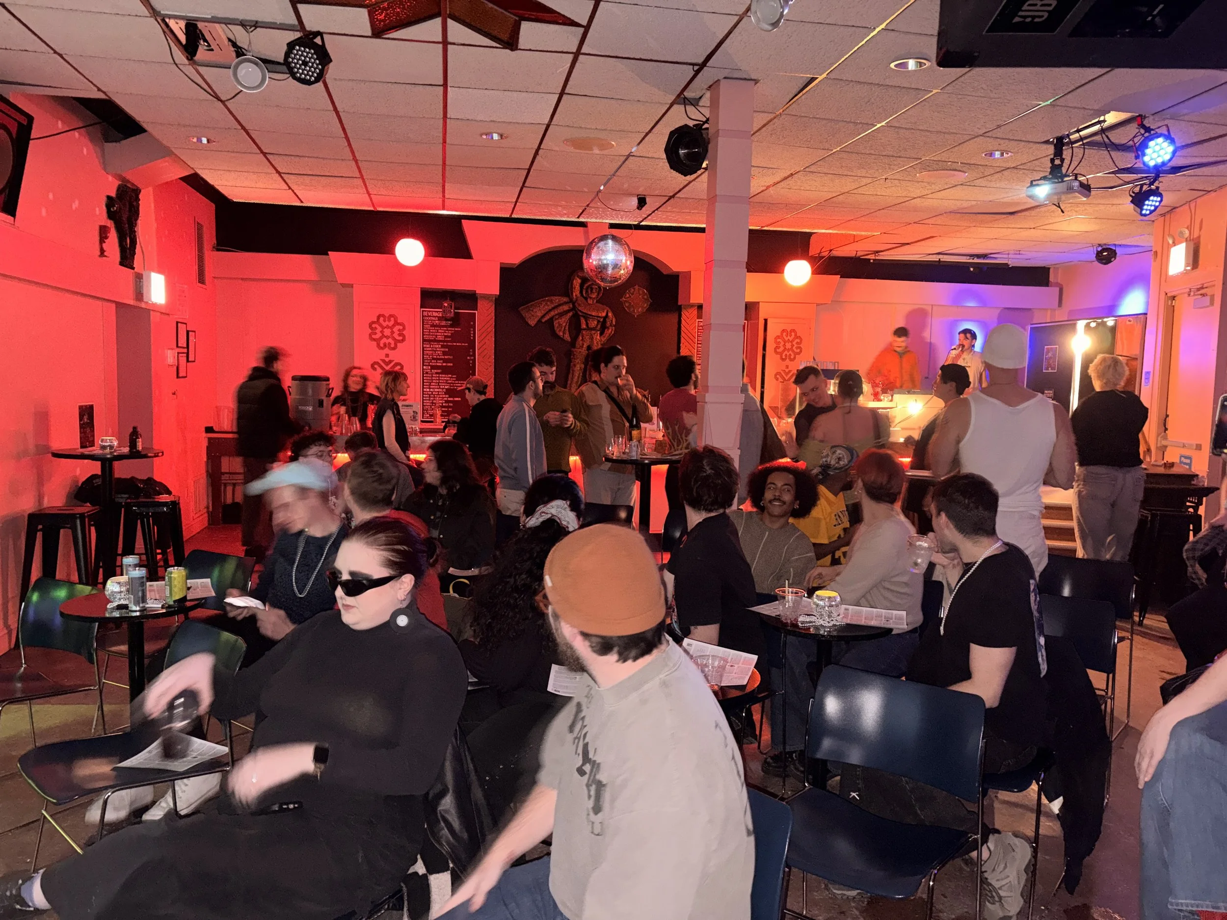 People gathering in a dimly lit bar or club with a stage and DJ booth, colorful lighting, and patrons sitting and standing around tables.