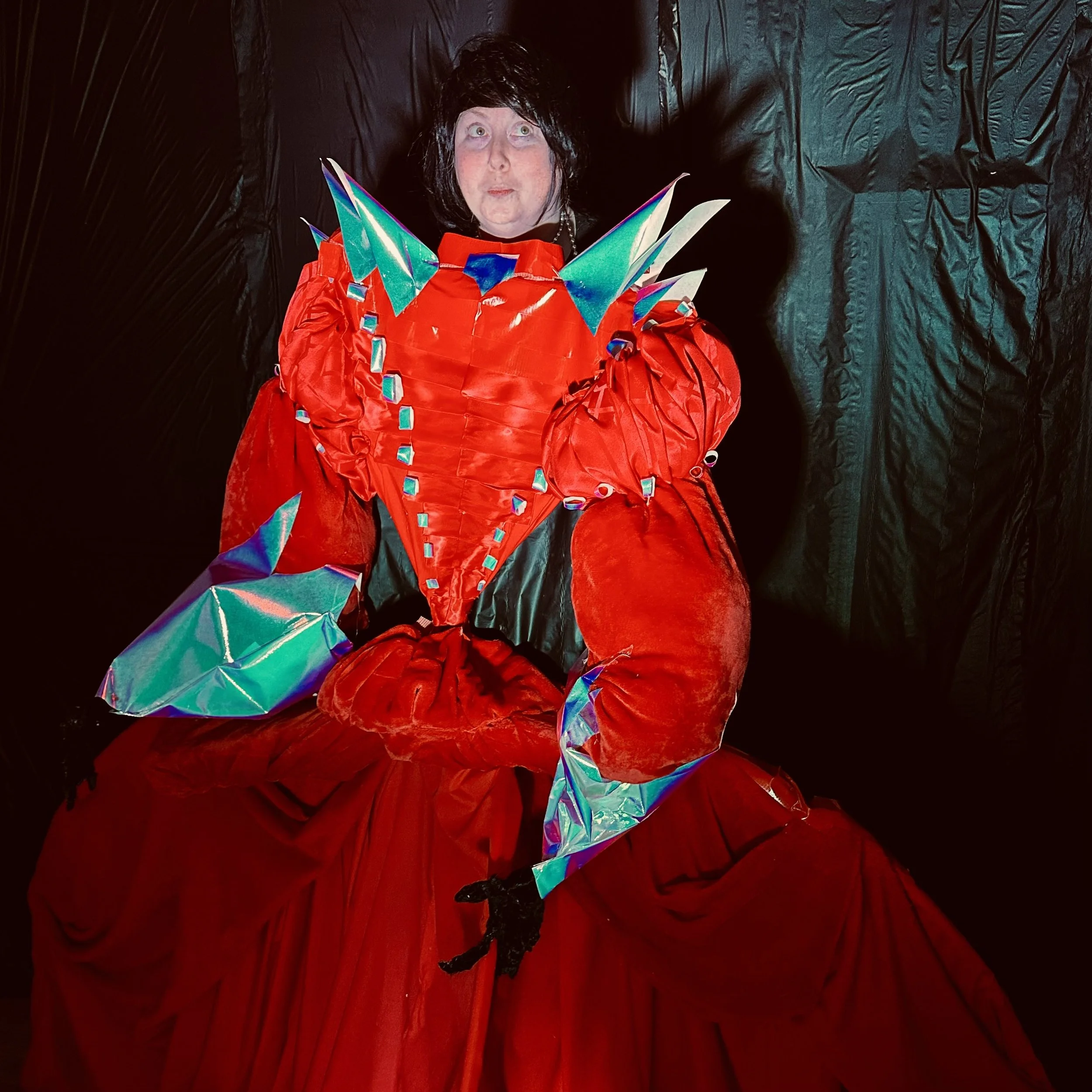 Person in elaborate red costume with colorful, holographic accents and black gloves, standing against a black backdrop.