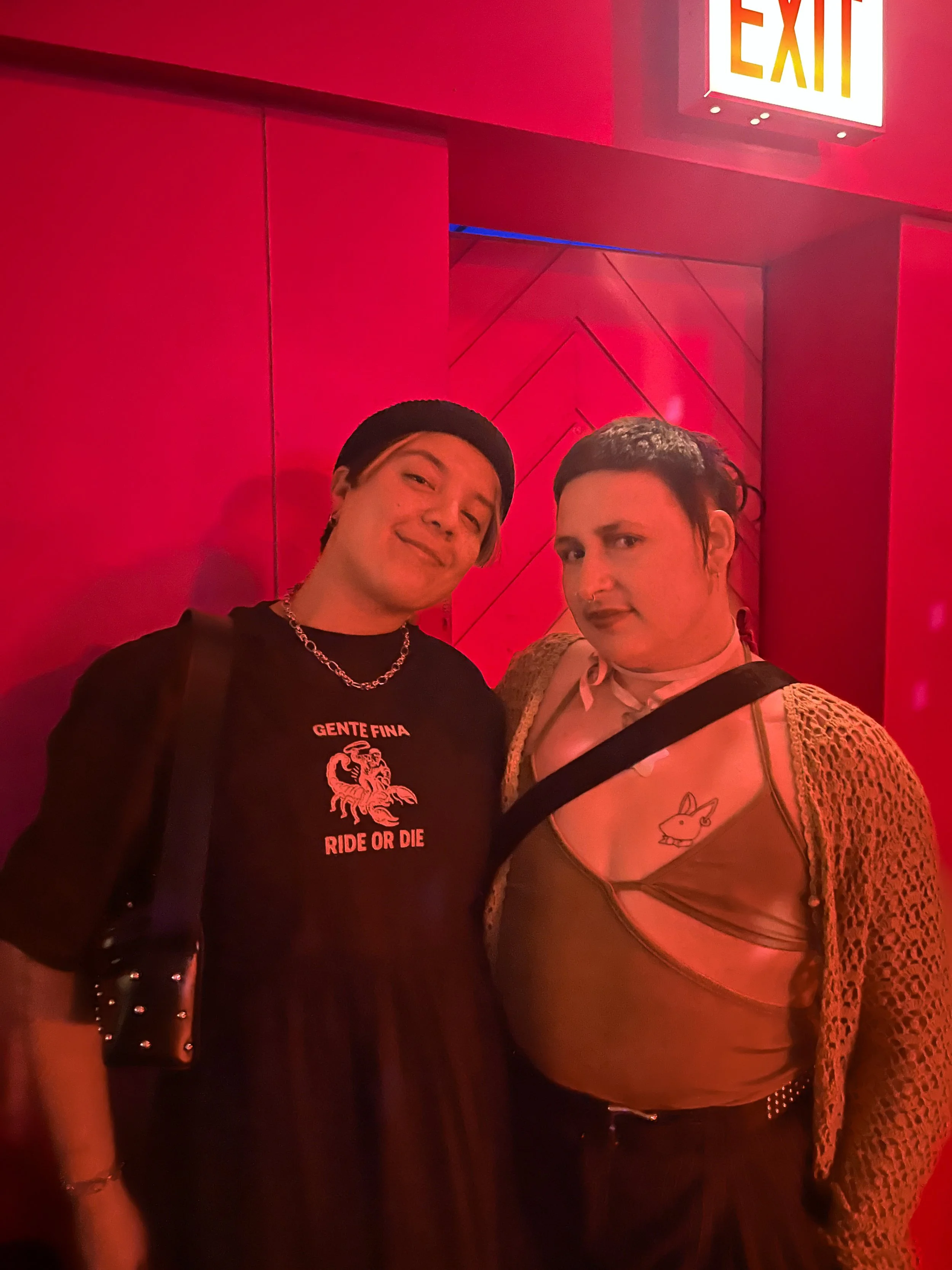 Two people posing in front of a red wall with an exit sign above them, wearing trendy clothing and makeup.