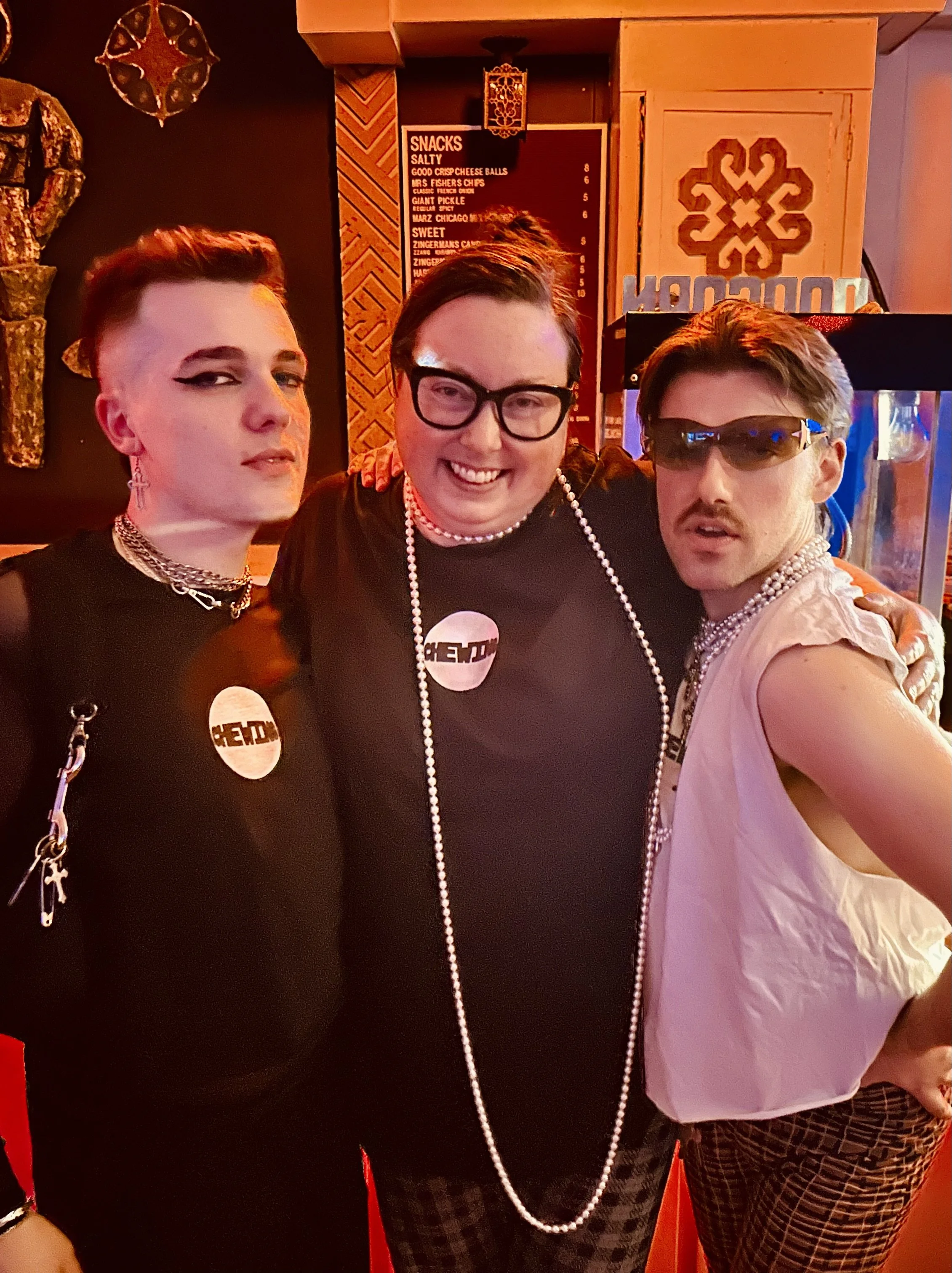 Three people dressed in 1980s inspired fashion posing together in a dimly lit bar or restaurant, with a snack menu in the background.