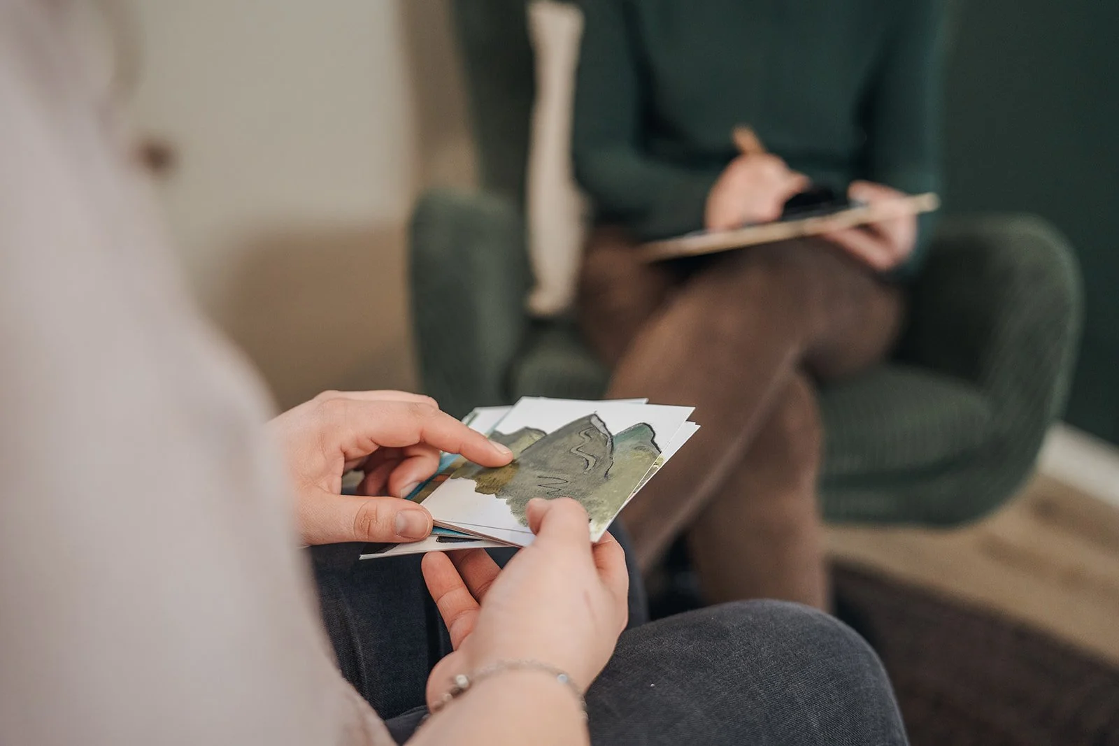 Zwei Personen sitzen in einem Raum, eine Person hält Postkarten mit Landschaftsbildern in den Händen, die andere Person sitzt in einem Sessel und schreibt auf einem Klemmbrett.