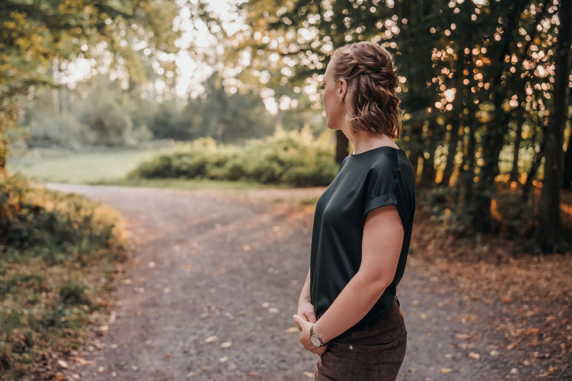 Eine Frau steht seitlich in einem Waldweg, umgeben von Bäumen und Herbstlaub, bei Sonnenuntergang.