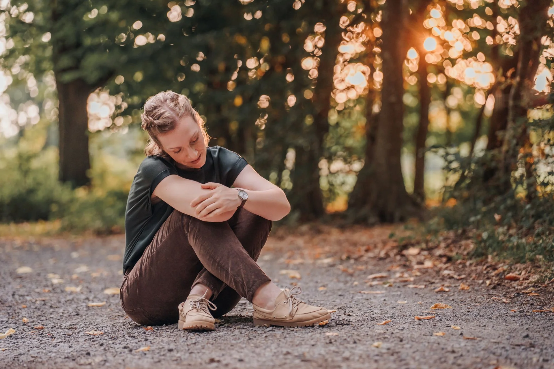 Junge Frau sitzt sich umarmend auf einem Weg im Wald bei Sonnenuntergang, umgeben von Bäumen und gefallenen Blättern.