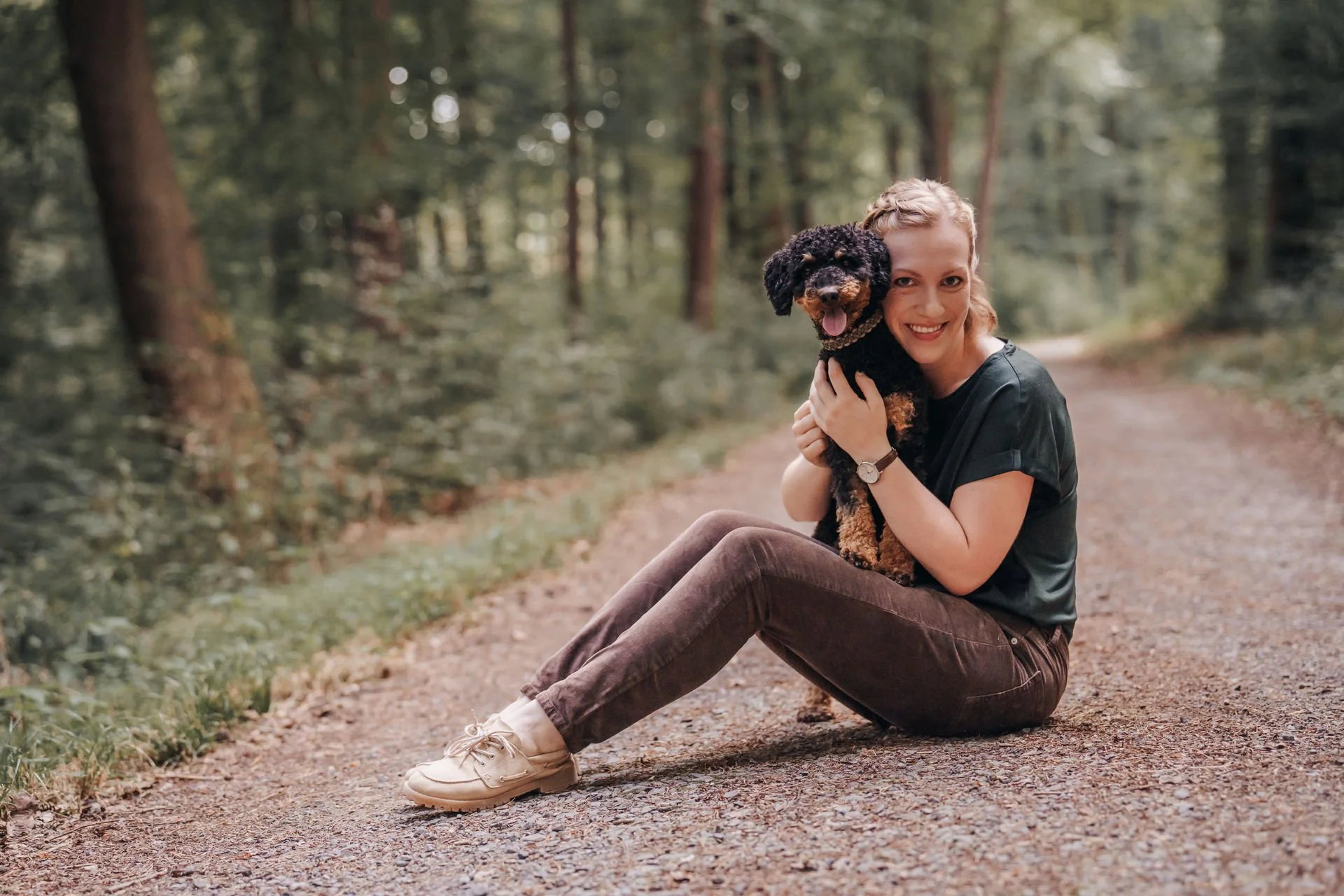Eine lachende Frau sitzt auf einem Waldweg, hält einen schwarzen und braunen Hund in den Armen.